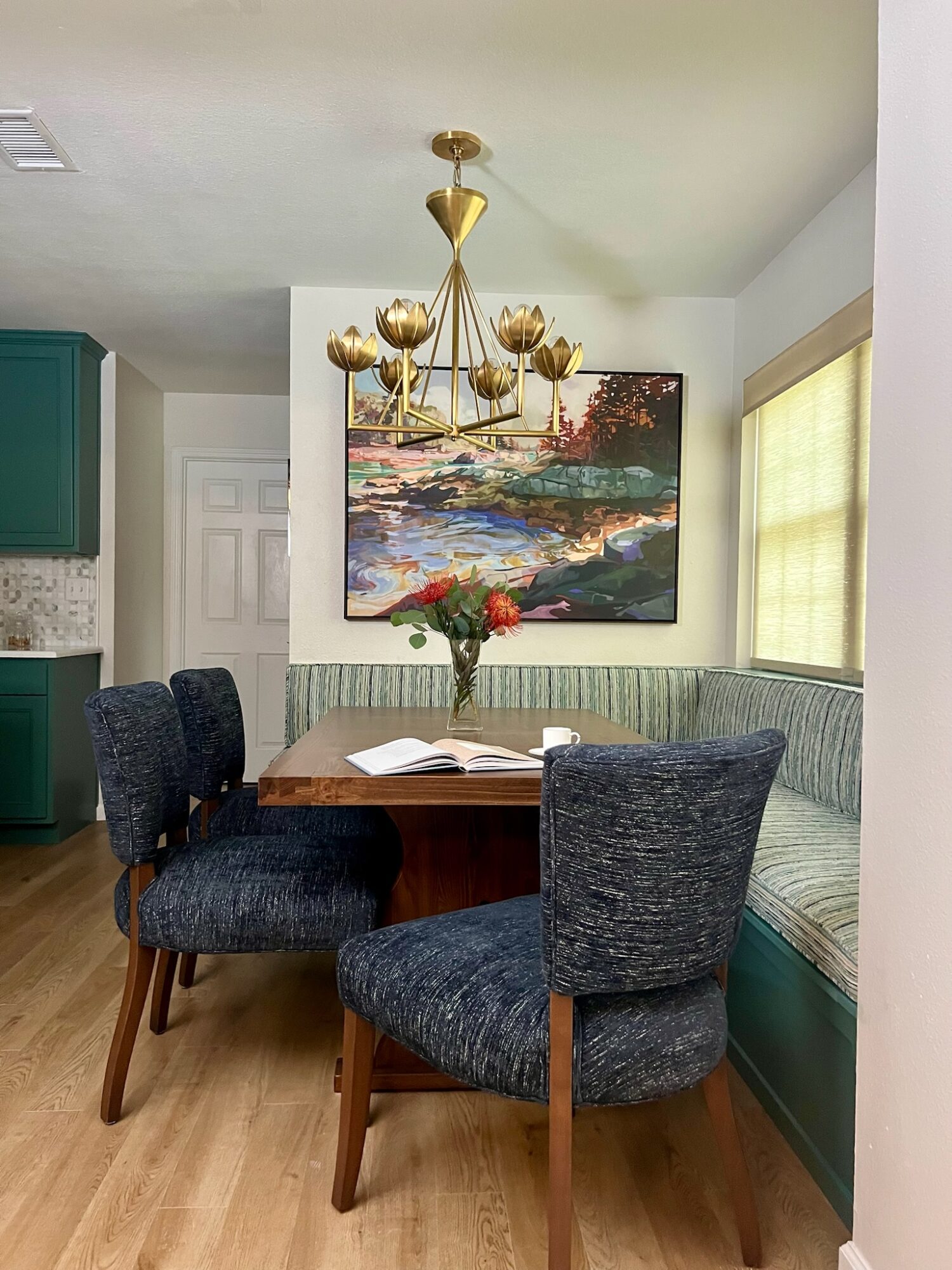Dining area with a round wooden table, four dark upholstered chairs, a floral centerpiece, a large landscape painting, and a window with blinds.