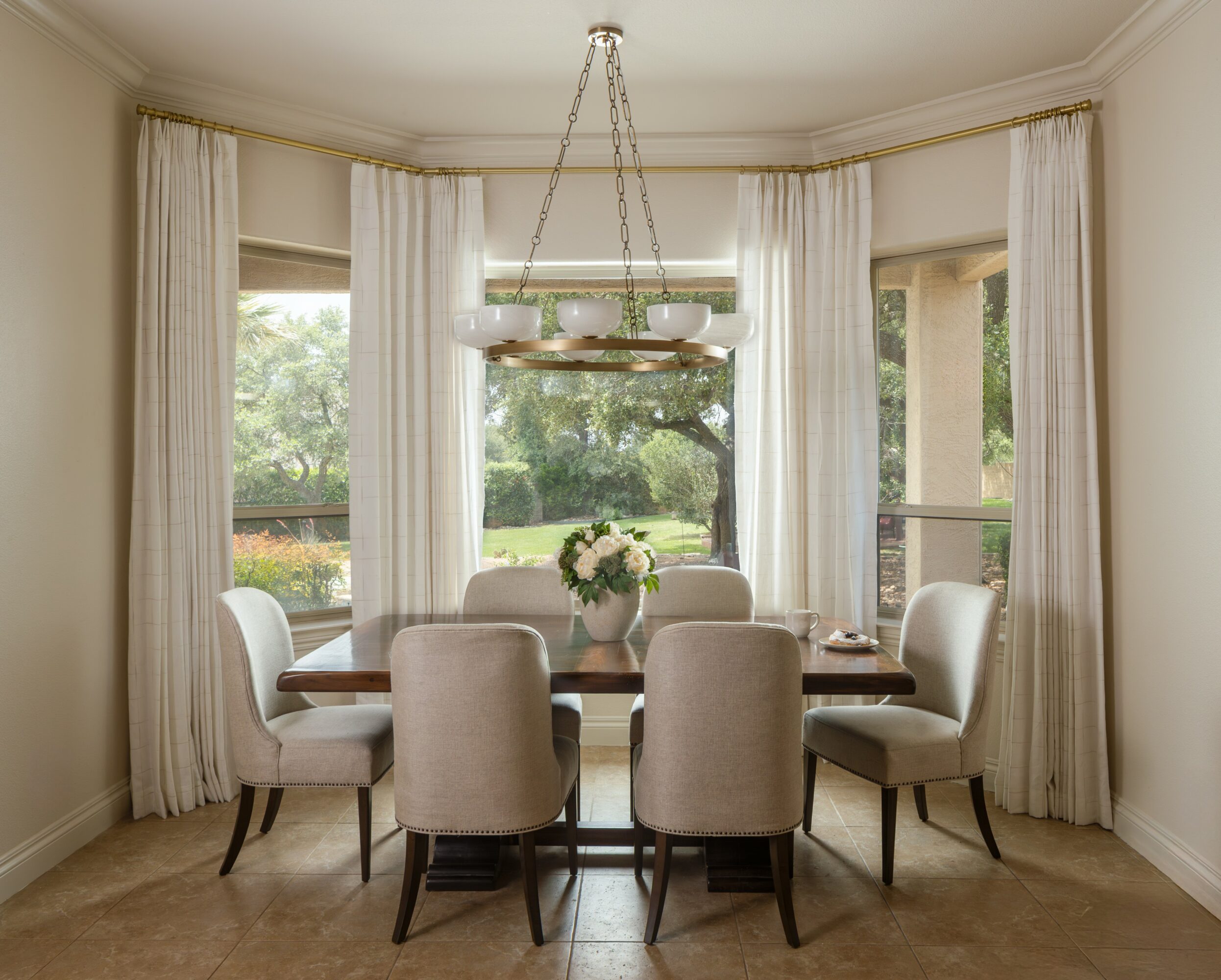 Dining room with a wooden table, six upholstered chairs, a chandelier, and large windows with curtains, overlooking a garden.