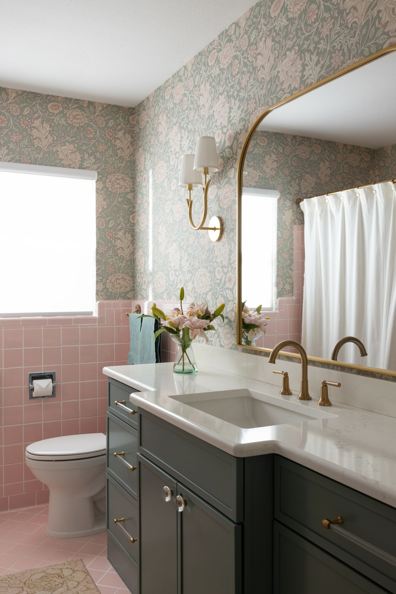 Bathroom with floral wallpaper, pink tiles, a mirror, a sink with gold fixtures, and a window with a curtain.
