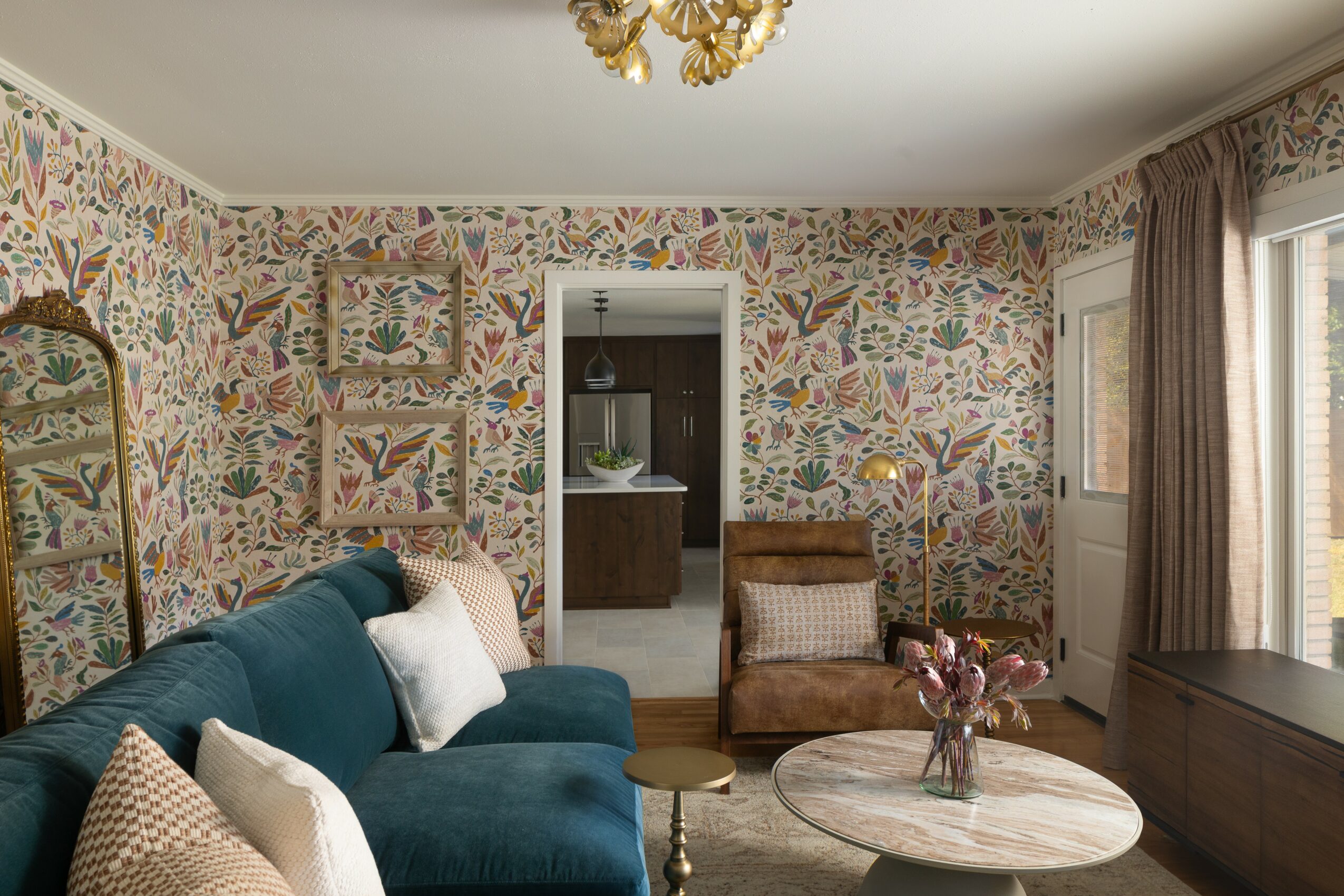 Living room with patterned wallpaper, a teal sofa with pillows, a round coffee table with flowers, and a window with curtains.