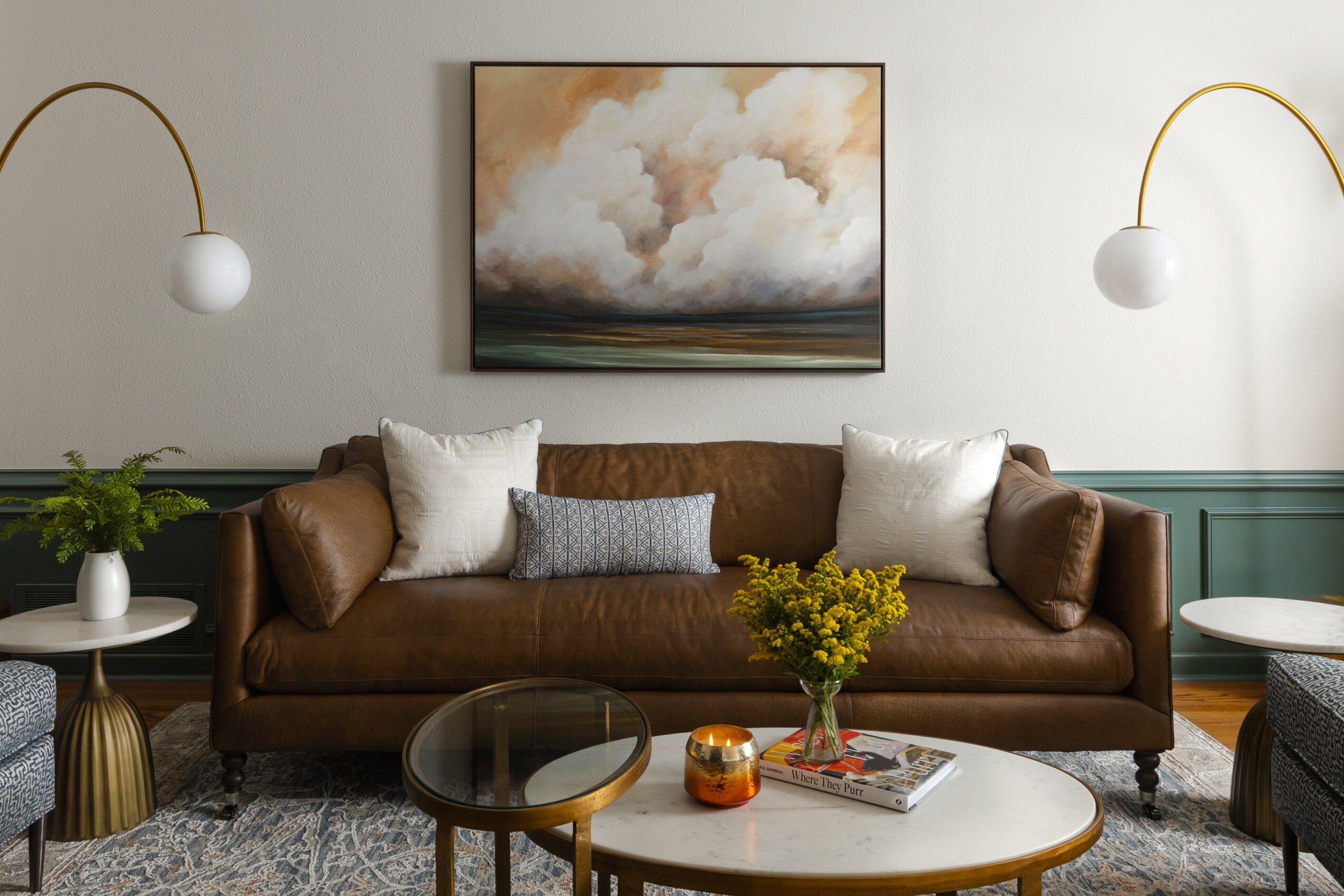 Living room with a brown sofa, white pillows, a painting, and two wall lamps, with a coffee table and decorative items.