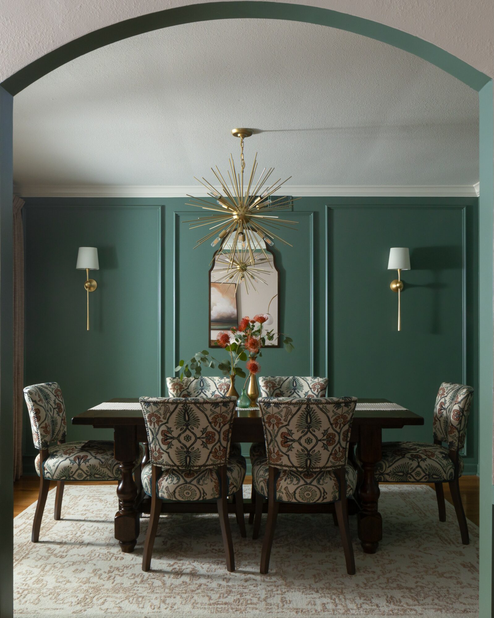 Dining room with green walls, six patterned chairs around a dark wooden table, a mirror, and wall sconces.