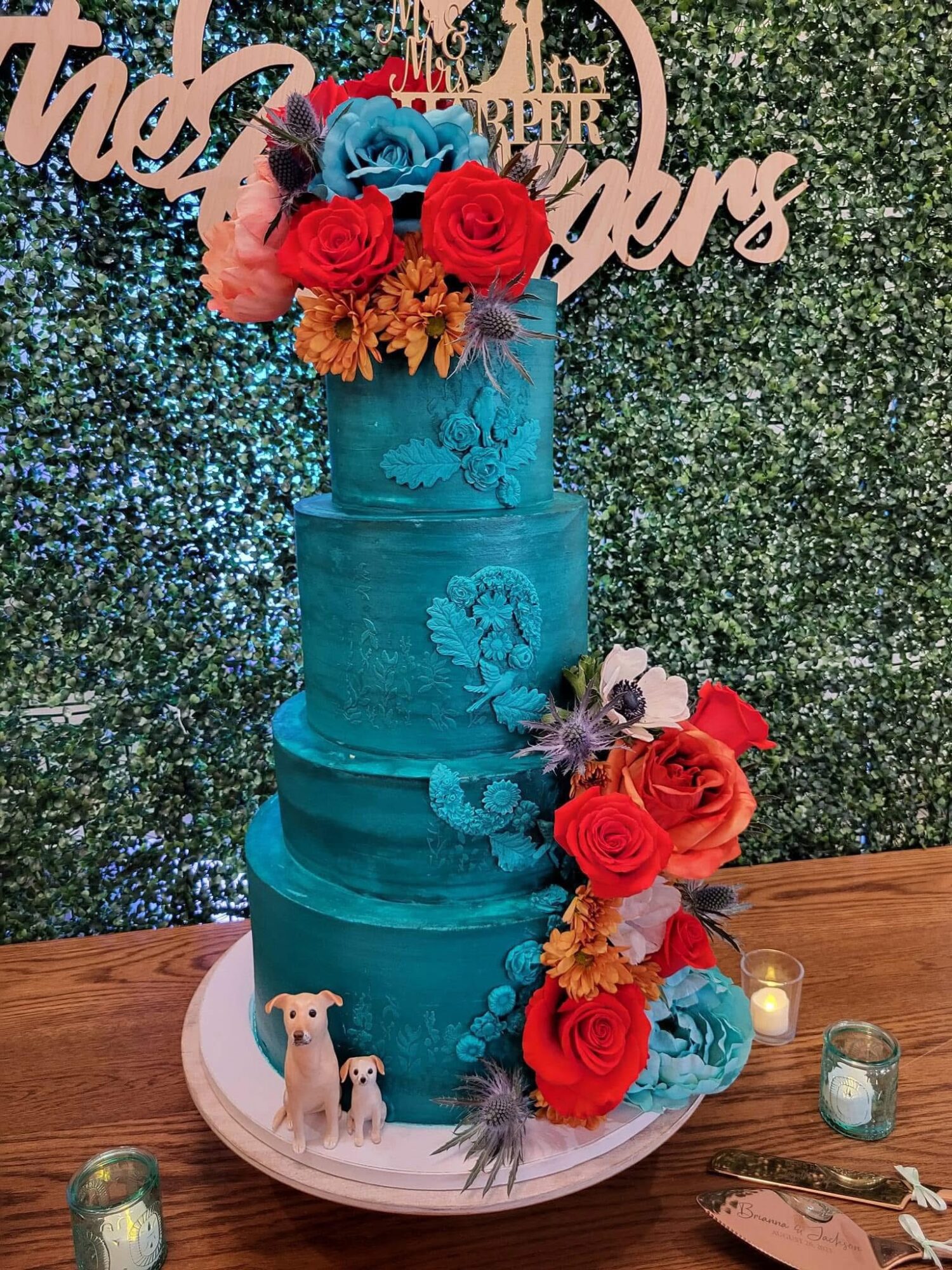 Four-tiered blue wedding cake decorated with red, pink, and orange flowers, with small dog figurines and candles nearby.