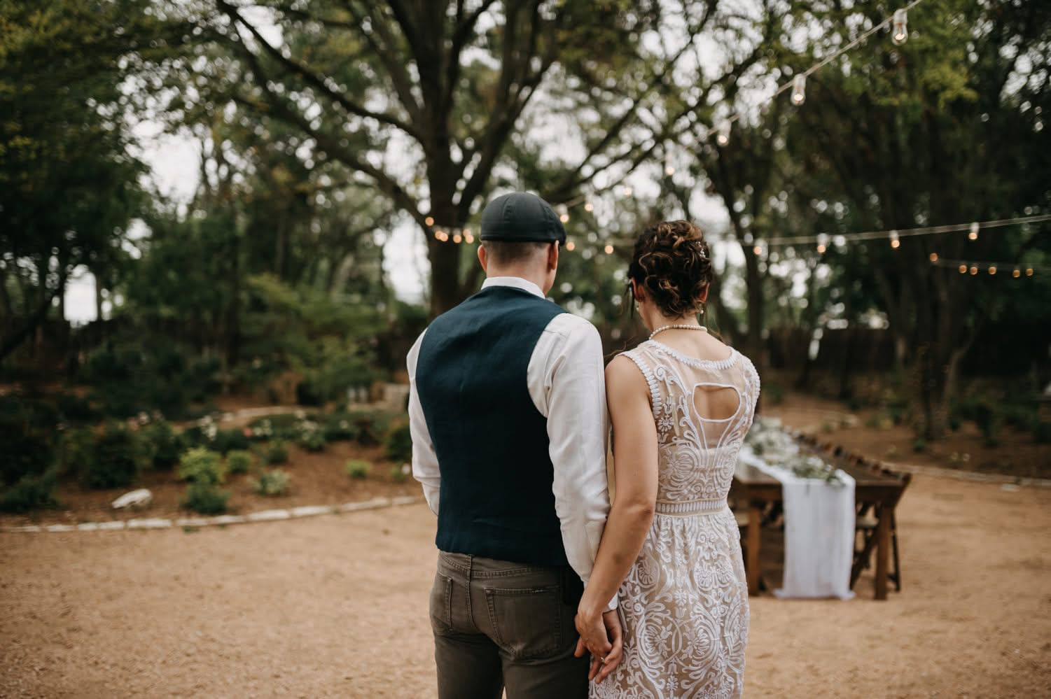 Two people stand outdoors, facing away, with trees and string lights in the background.