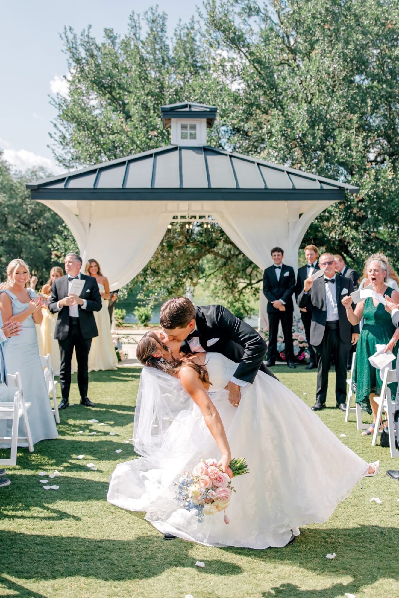 A bride and groom kiss under a gazebo at an outdoor wedding, surrounded by guests clapping and smiling.