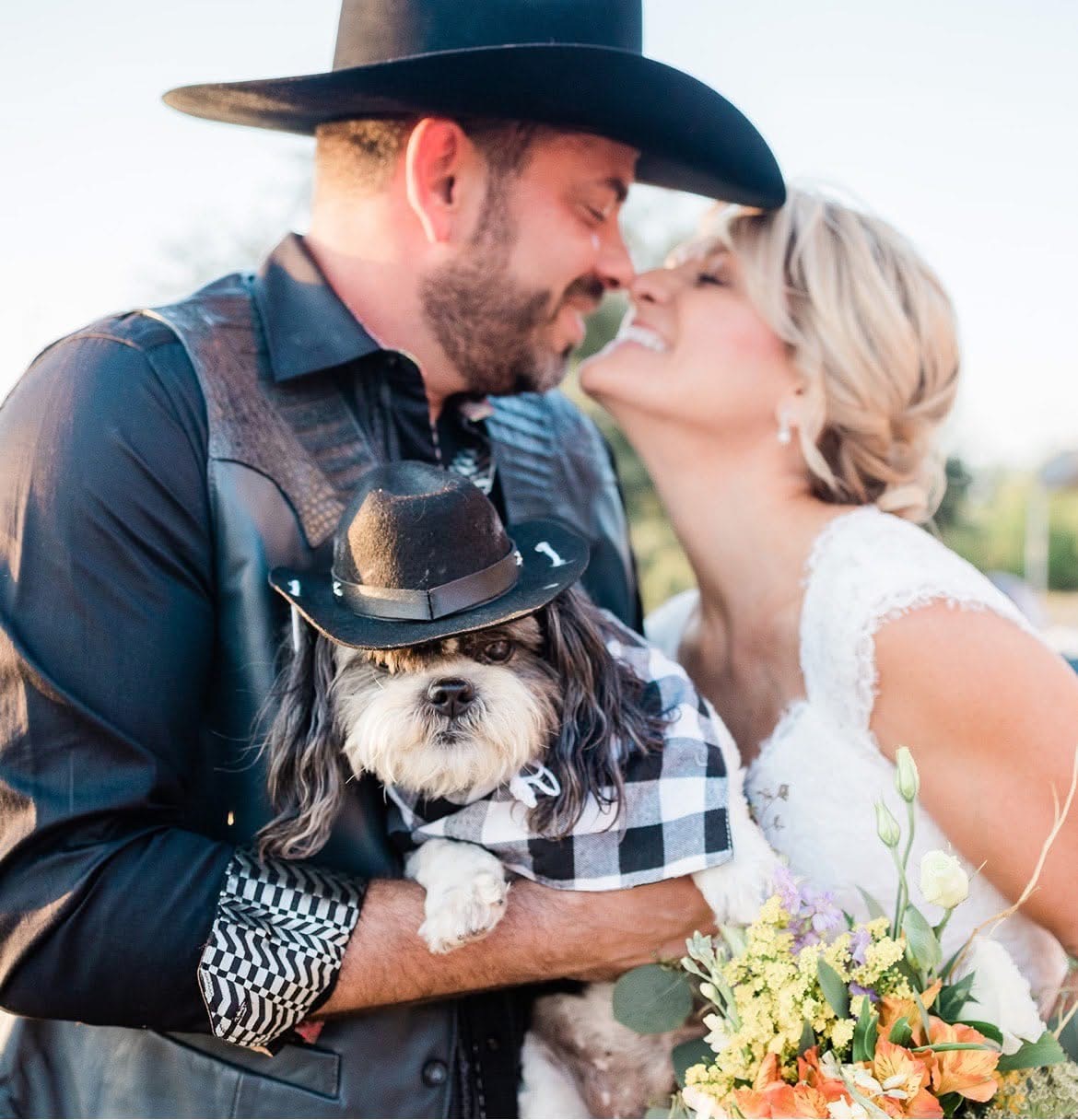 Man, woman, and dog outdoors, close-up, man wearing a hat, woman holding a bouquet, dog wearing a hat, smiling faces.