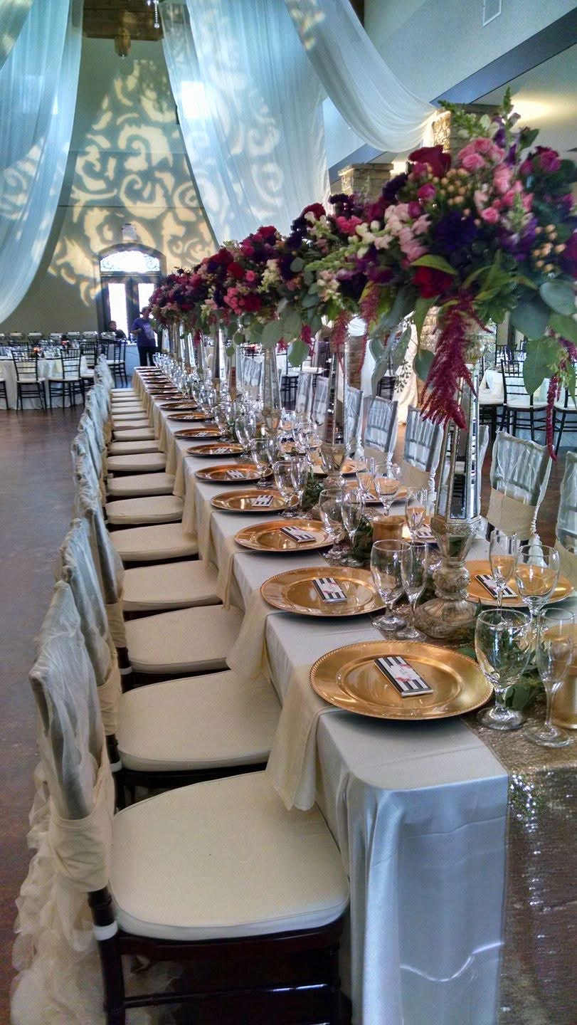 Long banquet table with floral centerpieces, set with plates, glasses, and napkins, under draped fabric and string lights.
