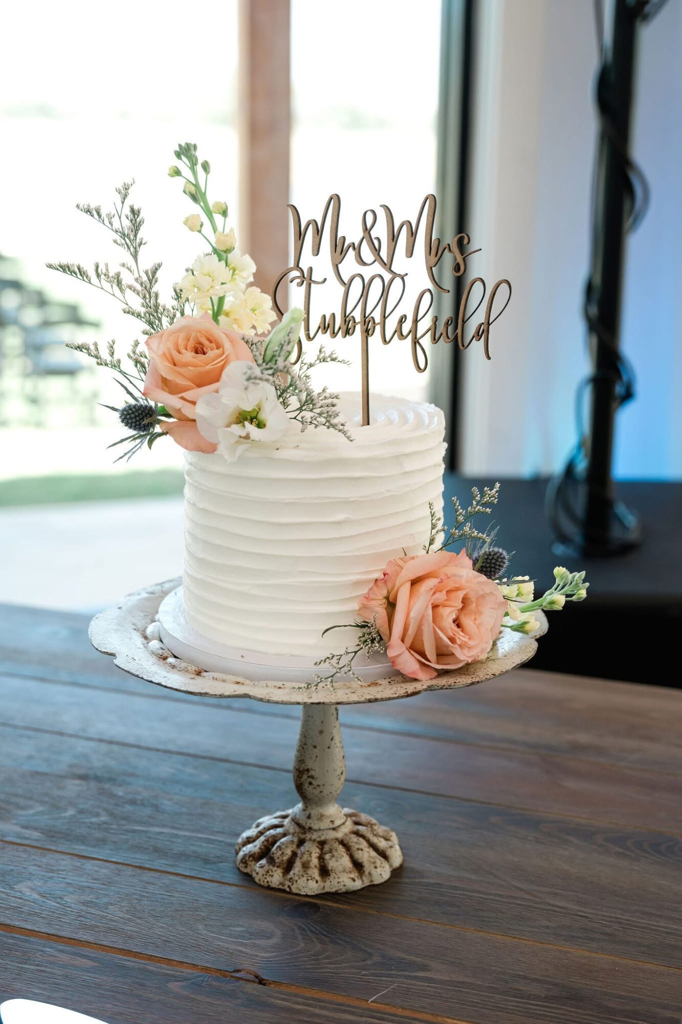 White wedding cake with pink and white flowers, on a decorative stand, with a cake topper reading