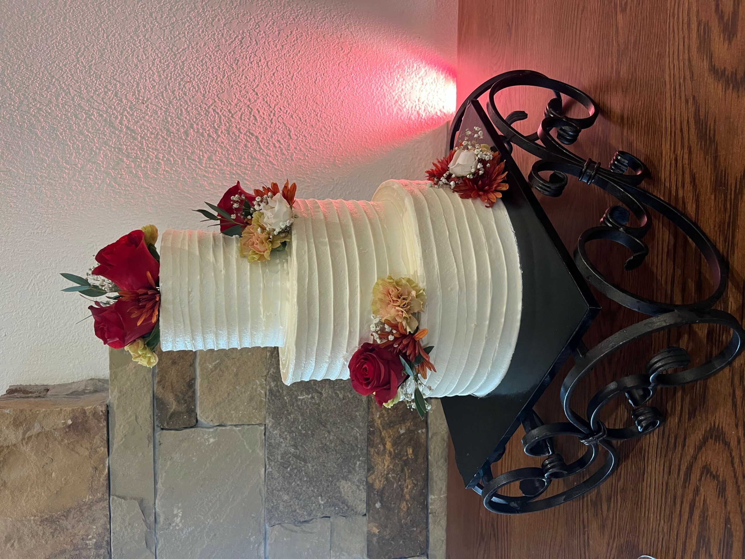 A multi-tiered white wedding cake decorated with red and white flowers, on a black stand with decorative scrolls, on a wooden surface.