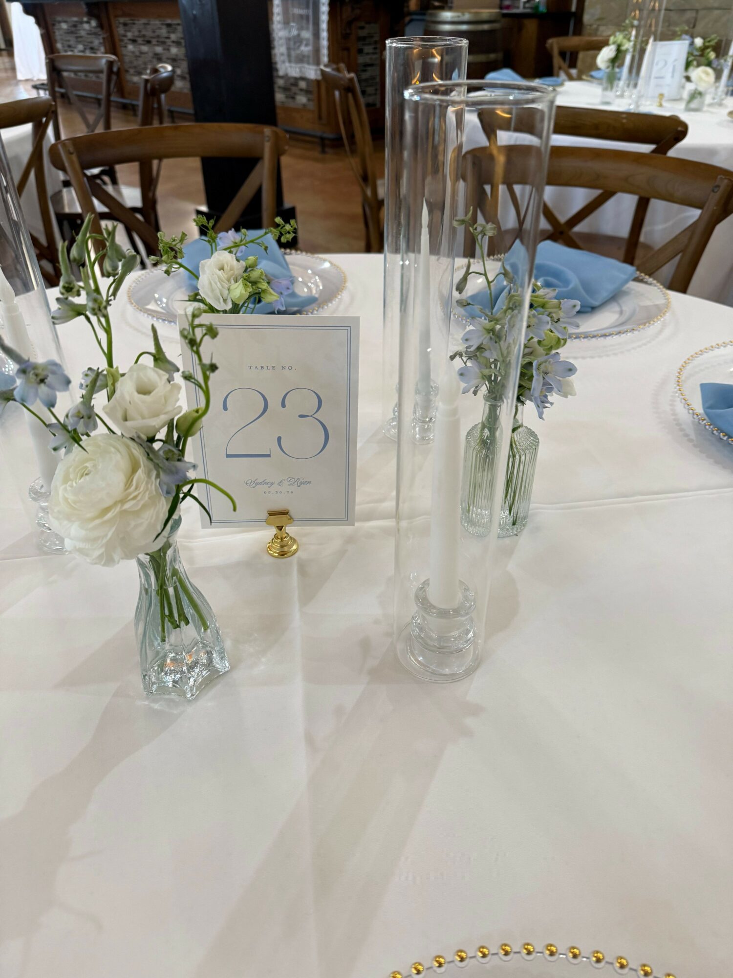 A table setting with a white flower in a glass vase, a card with the number 23, and a tall glass vase with flowers.