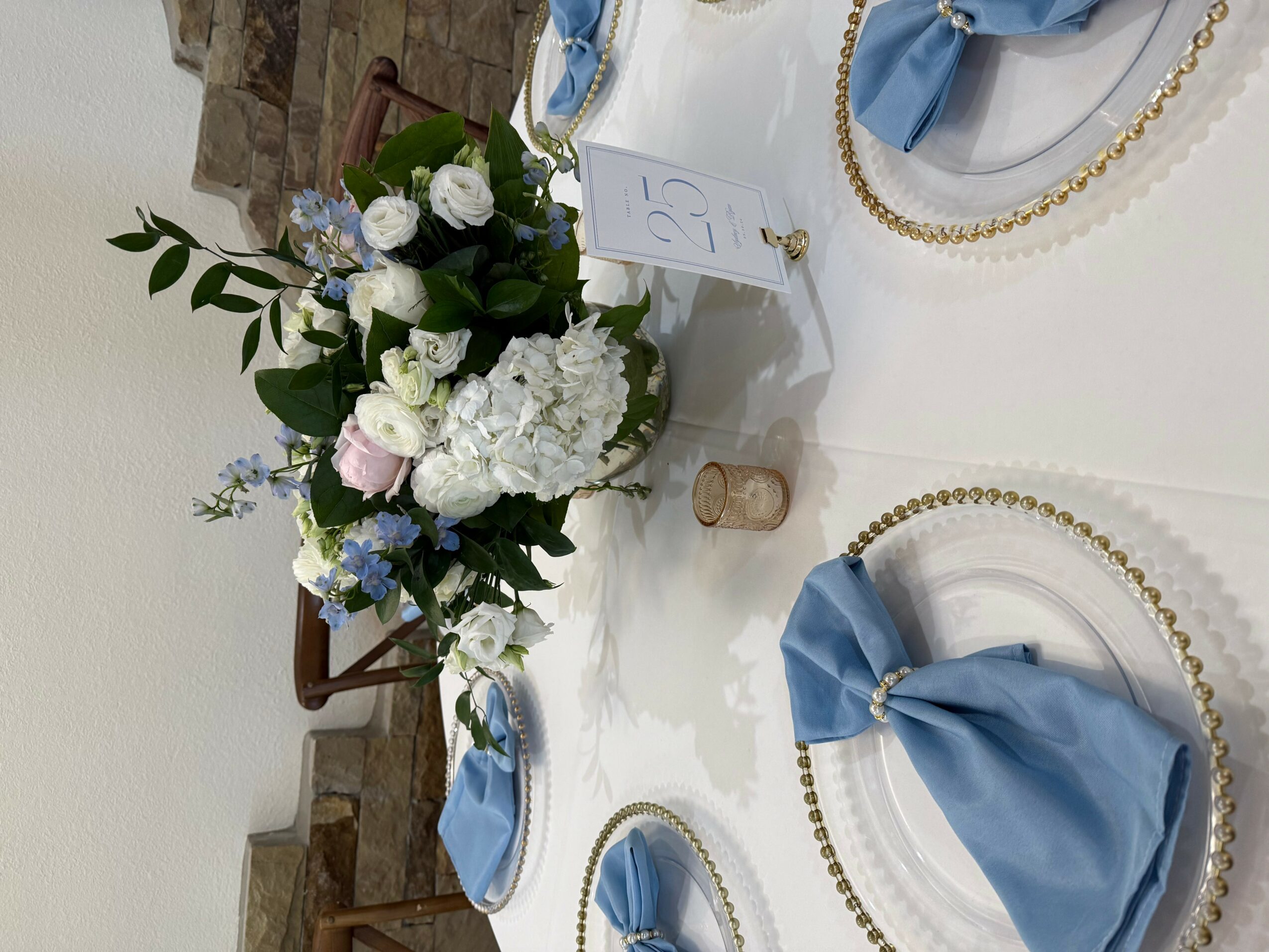 Table setting with floral centerpiece, blue napkins, gold beads, and a small card, viewed from the side.