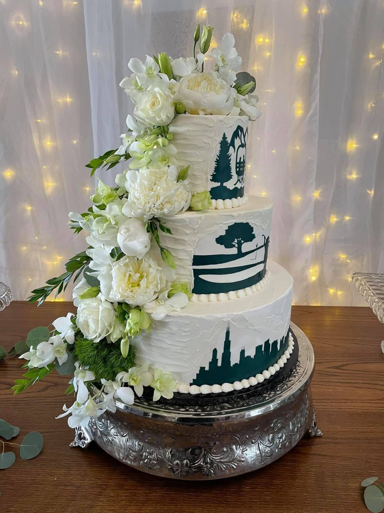 Three-tiered white wedding cake decorated with white flowers and green leaves, with silhouette designs on each tier, on a silver stand.
