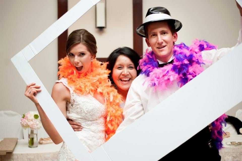 Three people smiling, wearing colorful feather boas, posing indoors with a white frame, one person in a hat.