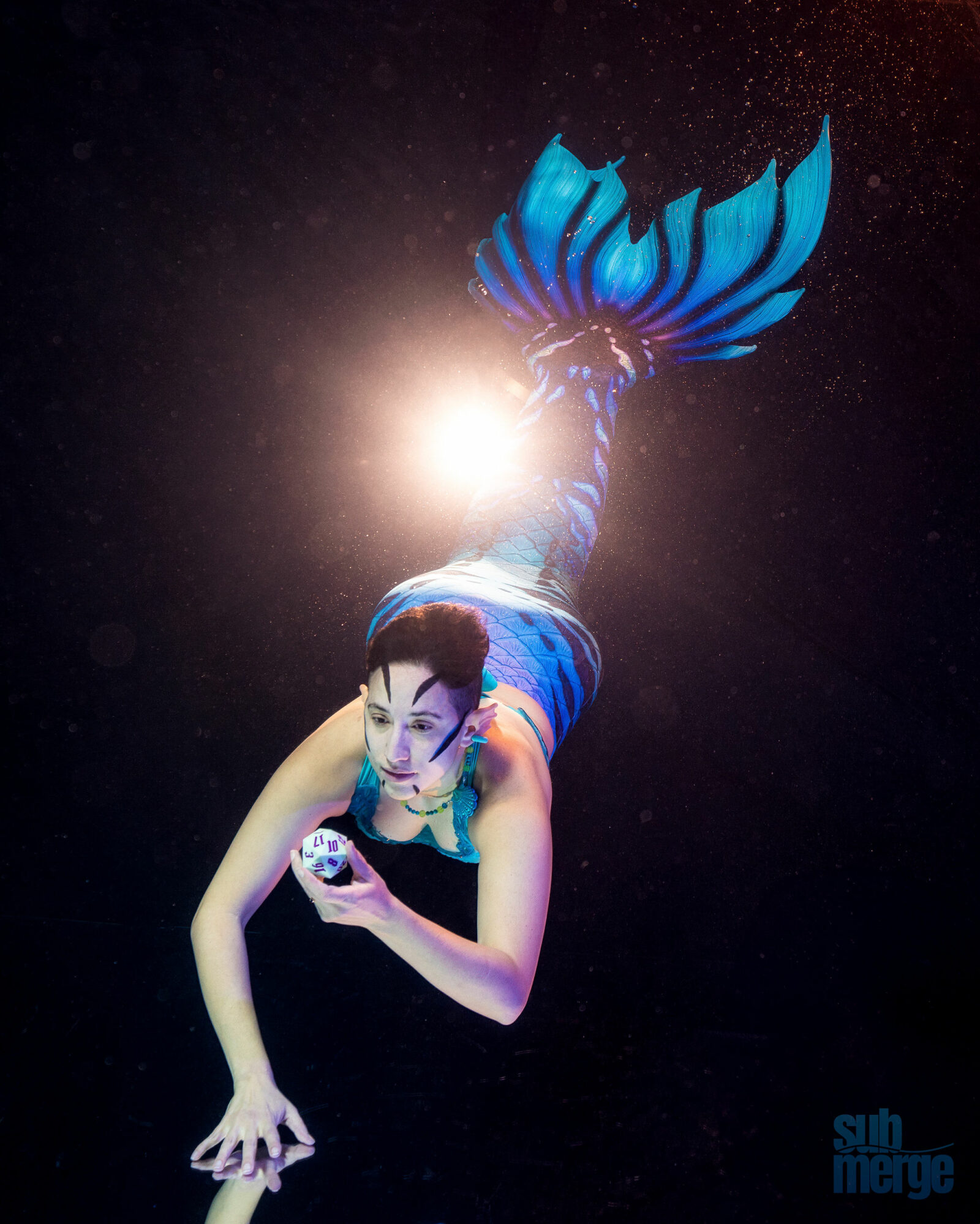 A person dressed as a mermaid with a blue tail floats underwater, holding a shiny object, with light shining behind them.