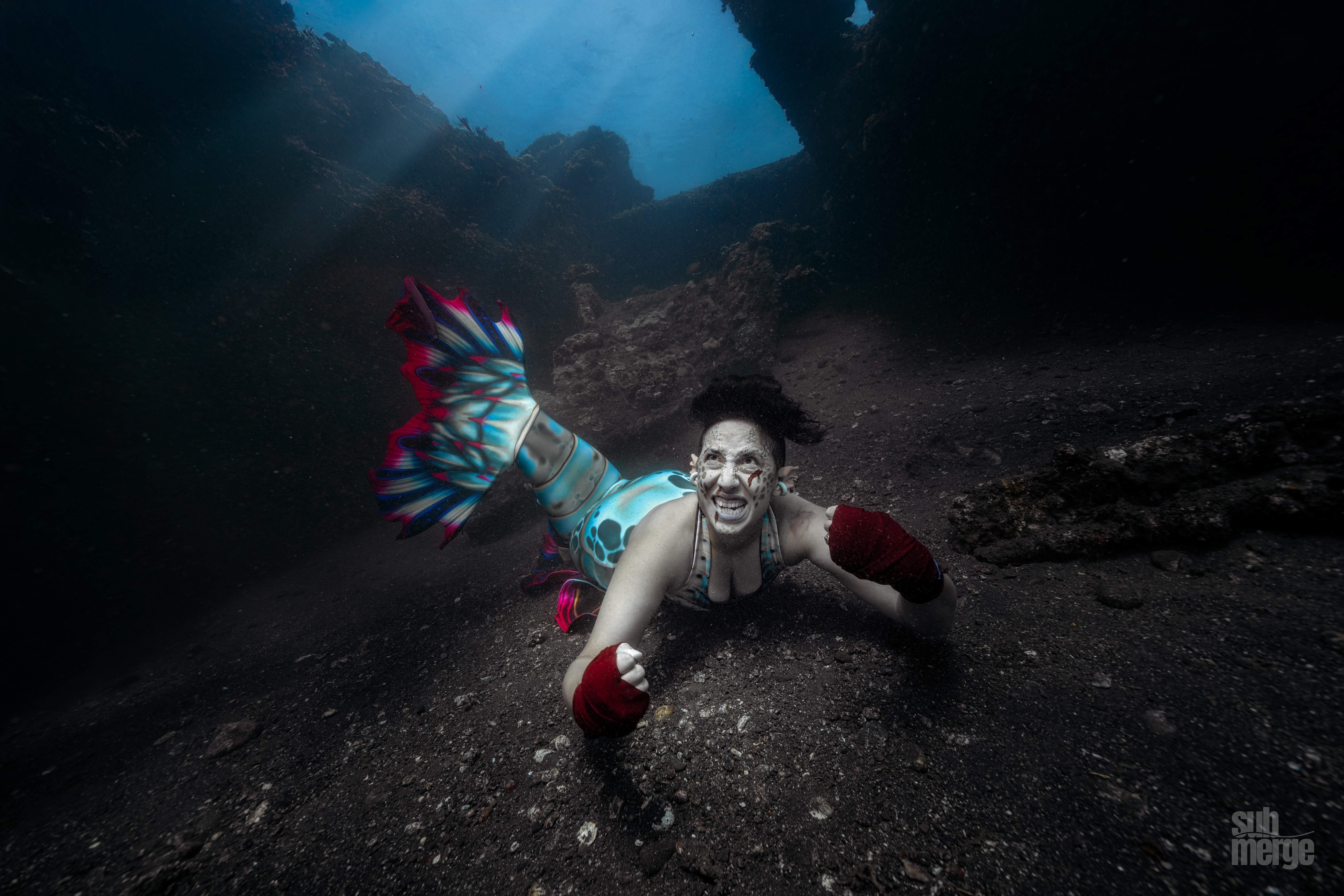 Person underwater smiling, wearing colorful fins and red gloves, with a dark underwater landscape in the background.