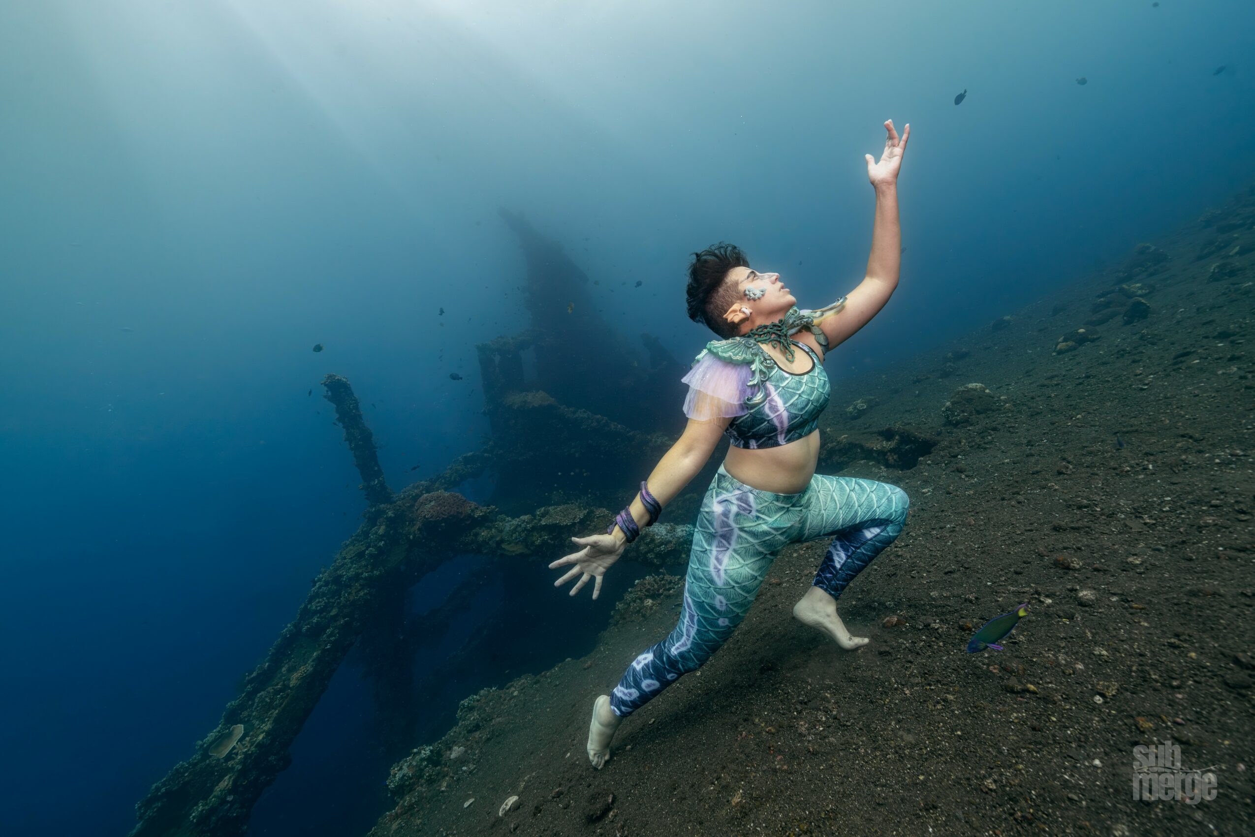 Person underwater near a sunken shipwreck, reaching upward with one arm, wearing a patterned outfit and mask.