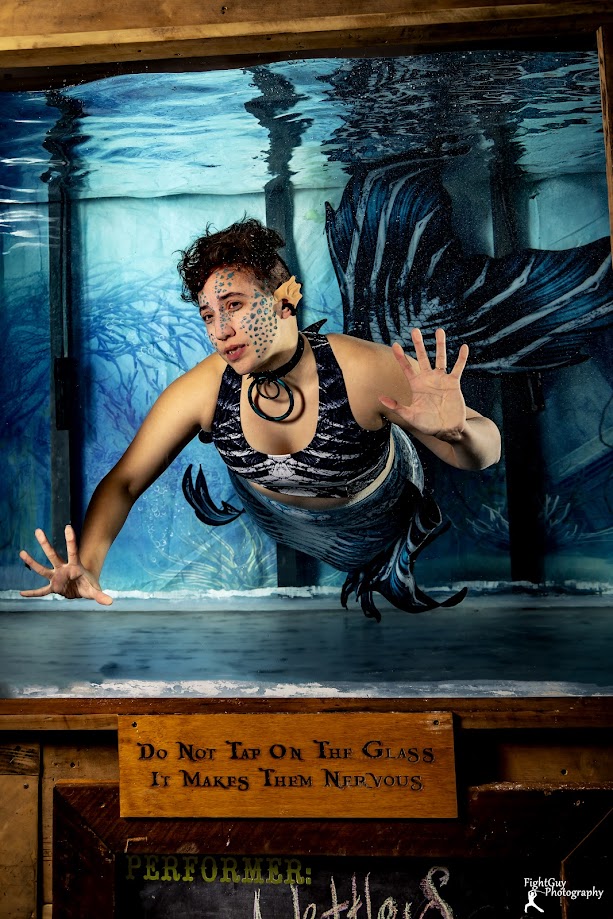 Person dressed as a mermaid underwater with face paint, reaching out with one hand, in front of an aquatic backdrop.