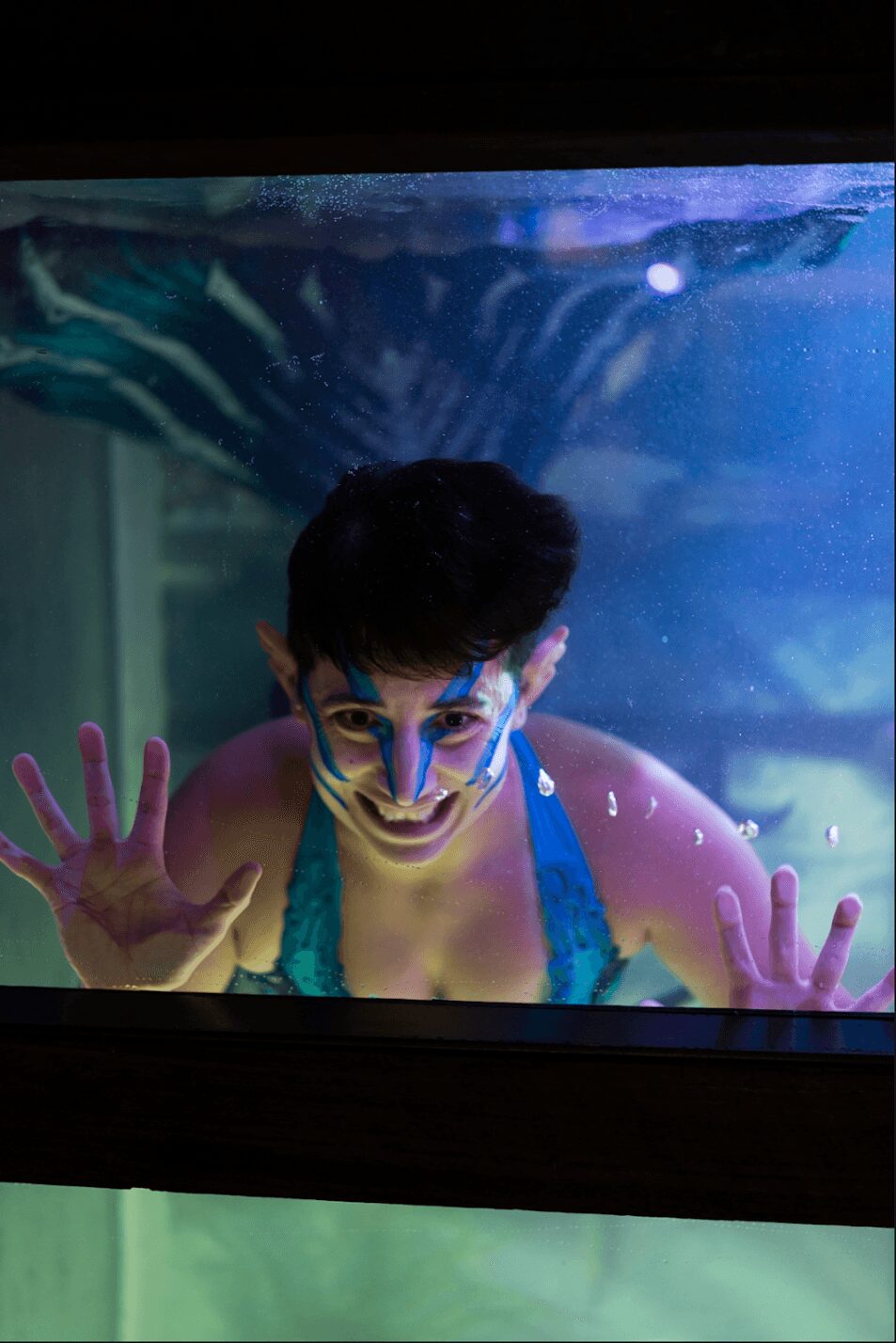 Person underwater wearing goggles, reaching towards the glass, with colorful background and water bubbles.