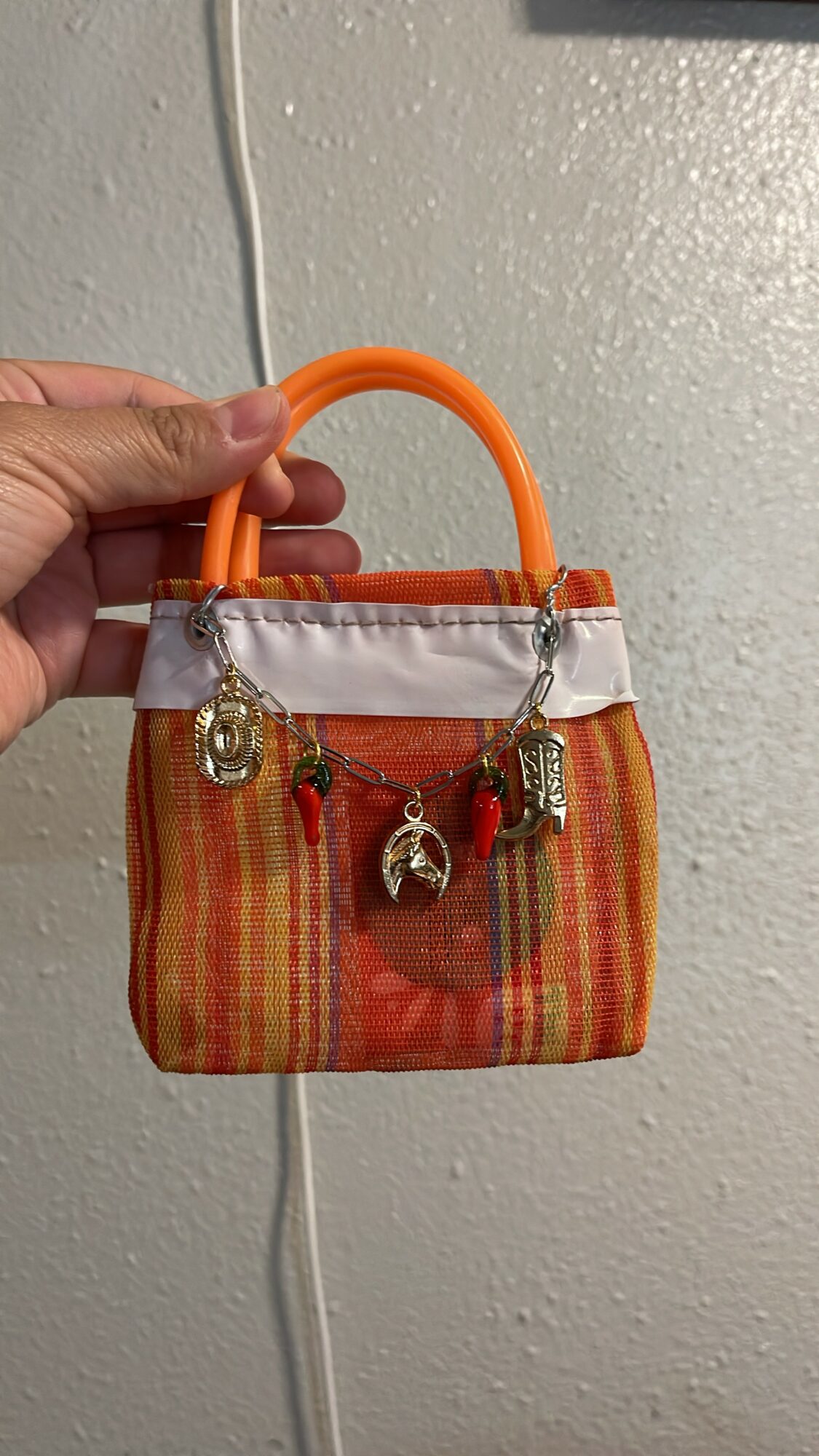 Small handbag with orange handle, striped fabric, and metal charms hanging from the front, held against a wall.