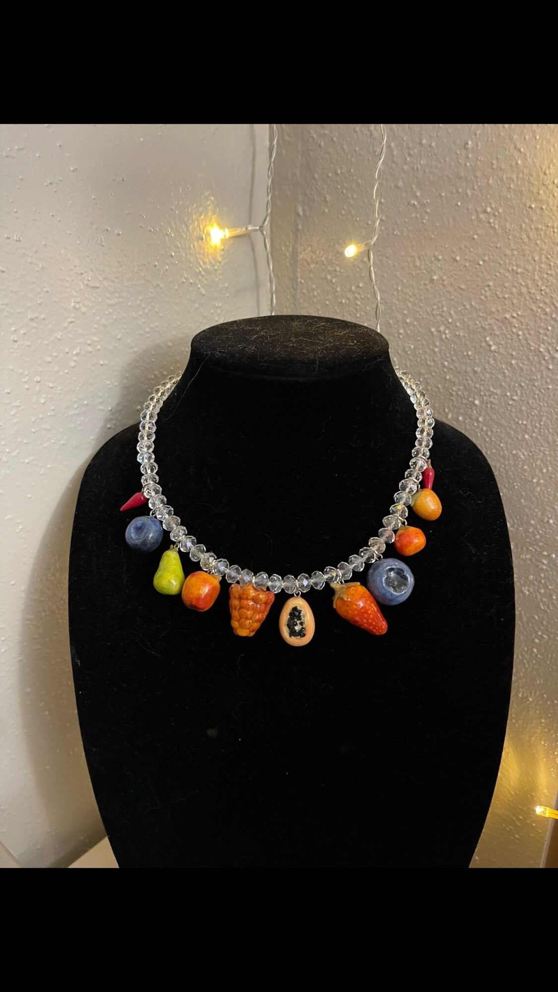 Necklace with colorful beads and a central pendant displayed on a black bust, against a light background with small lights.