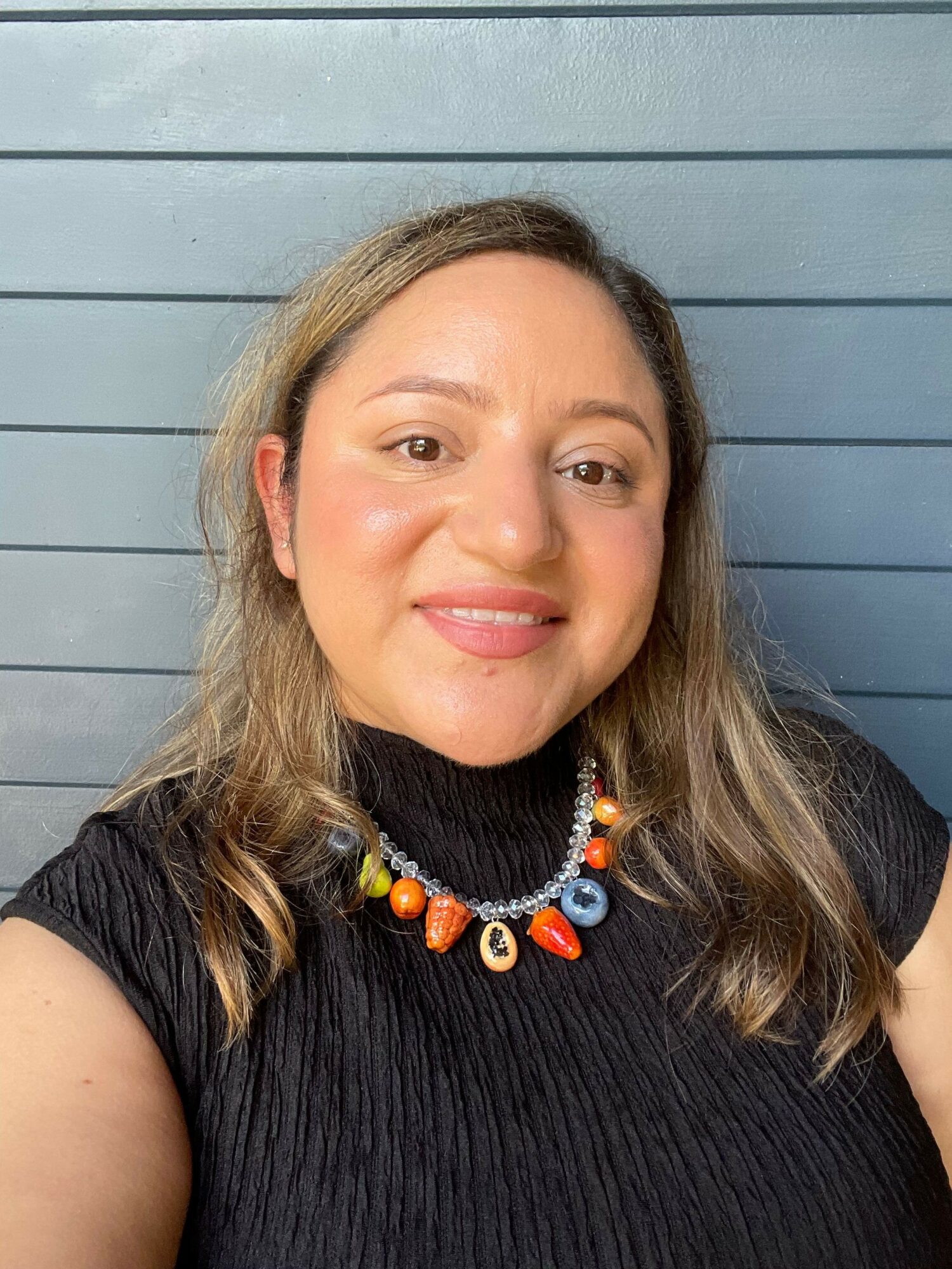 Woman with wavy hair wearing a black top and colorful necklace, smiling, against a blue wooden background.