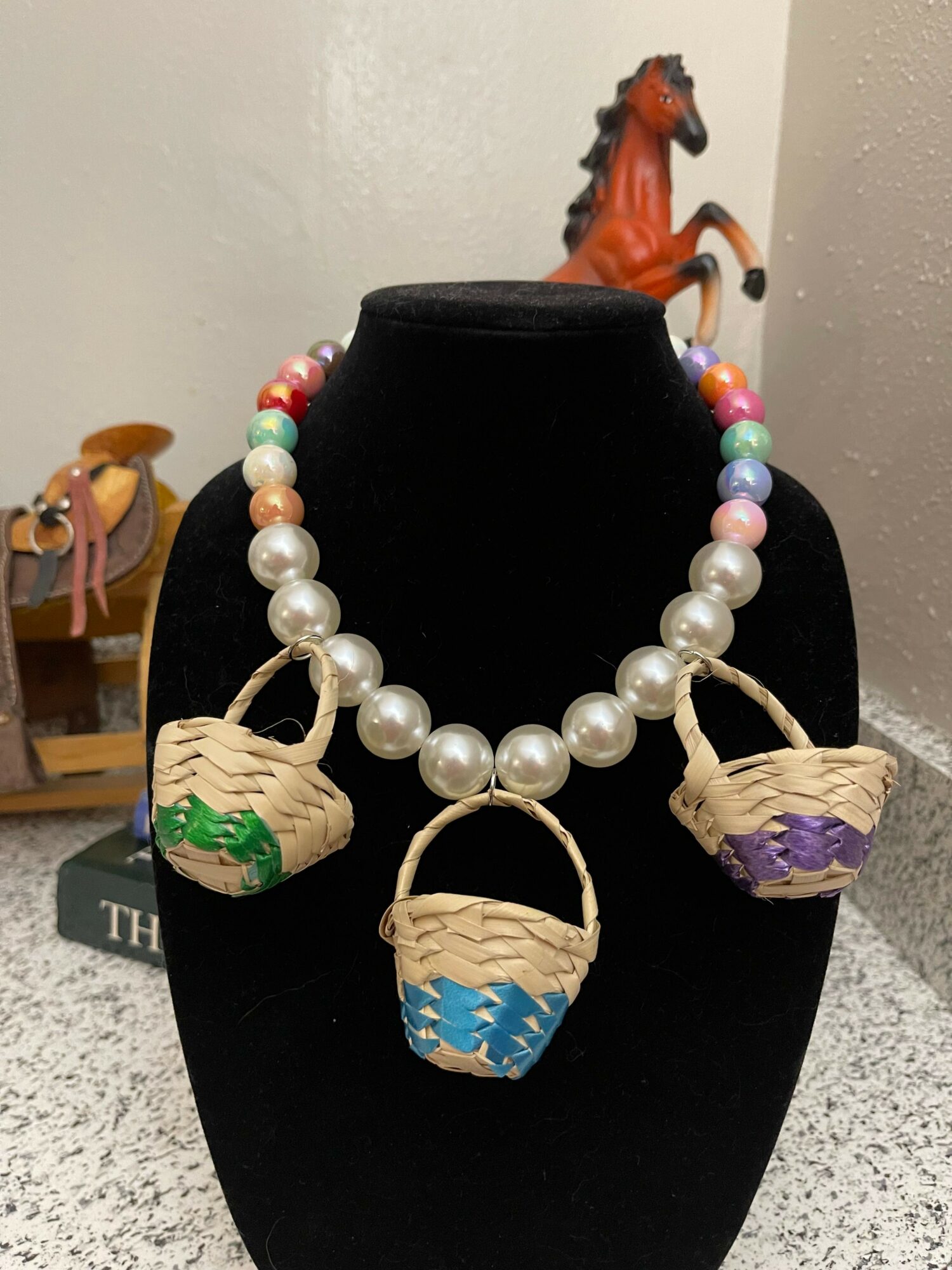 Pearl necklace with three small woven baskets hanging, each decorated with colorful patterns, displayed on a black stand.