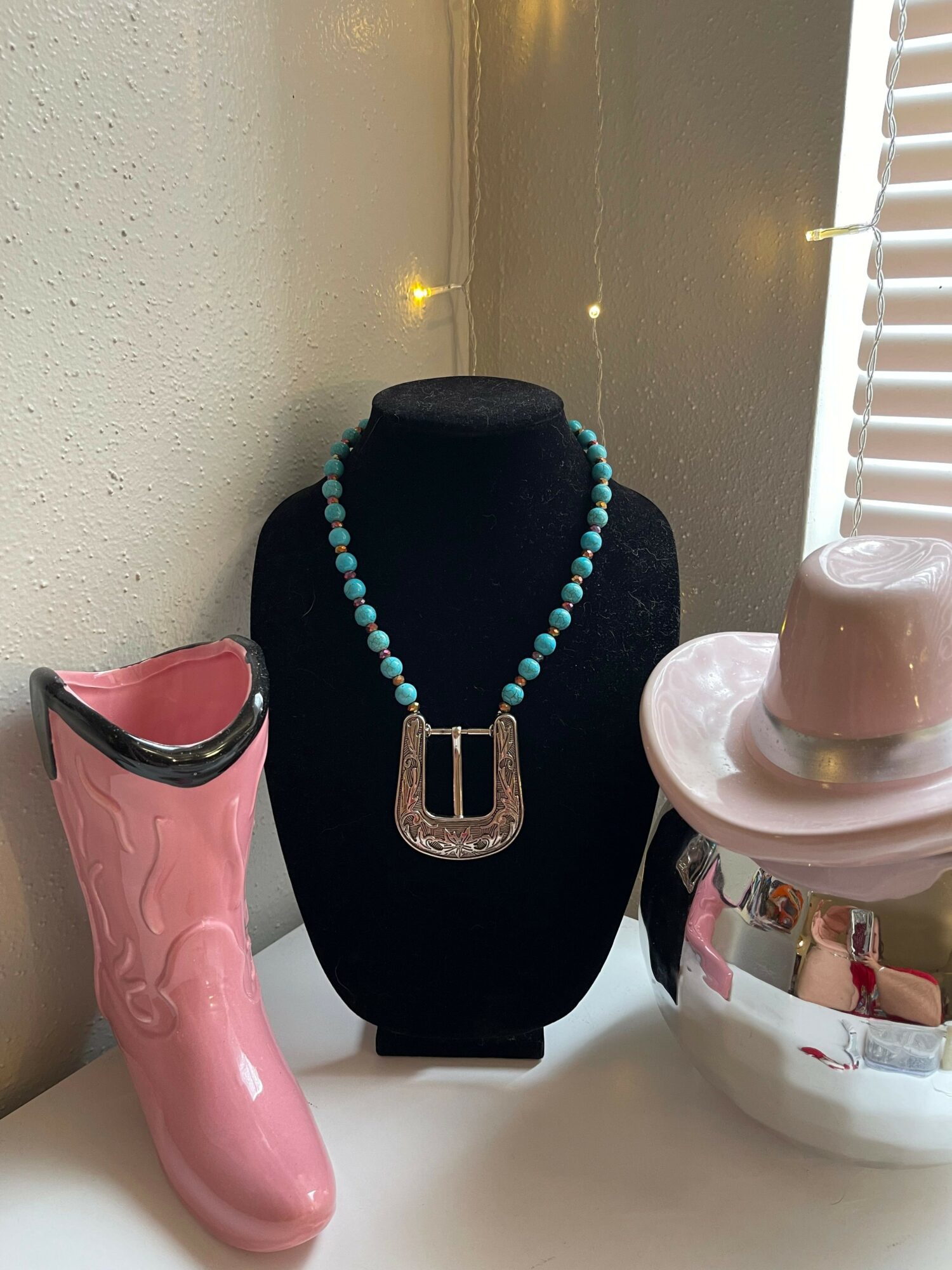 Necklace with turquoise beads and a silver buckle displayed on a black bust, with pink and white objects nearby.
