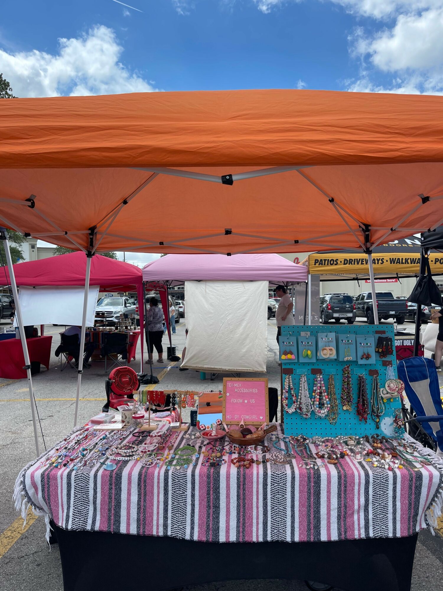 Outdoor market stall with a pink tablecloth, display of jewelry, and a person in the background under a blue sky.