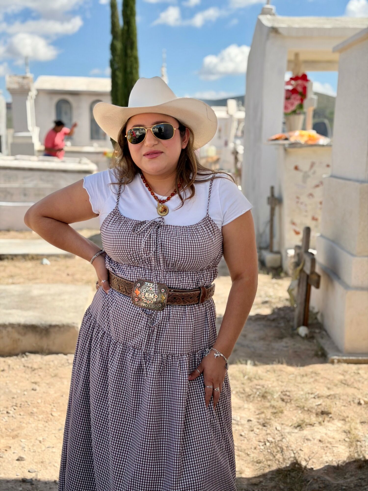 Woman wearing sunglasses, a wide-brimmed hat, and a patterned dress, standing outdoors with white structures and blue sky.