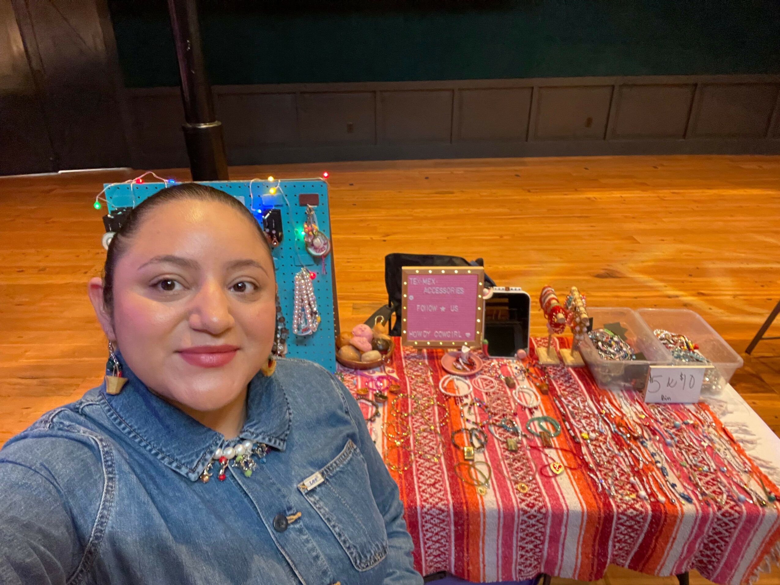 Person with short dark hair and red lipstick sitting at a table with colorful fabric and electronic components, facing camera.