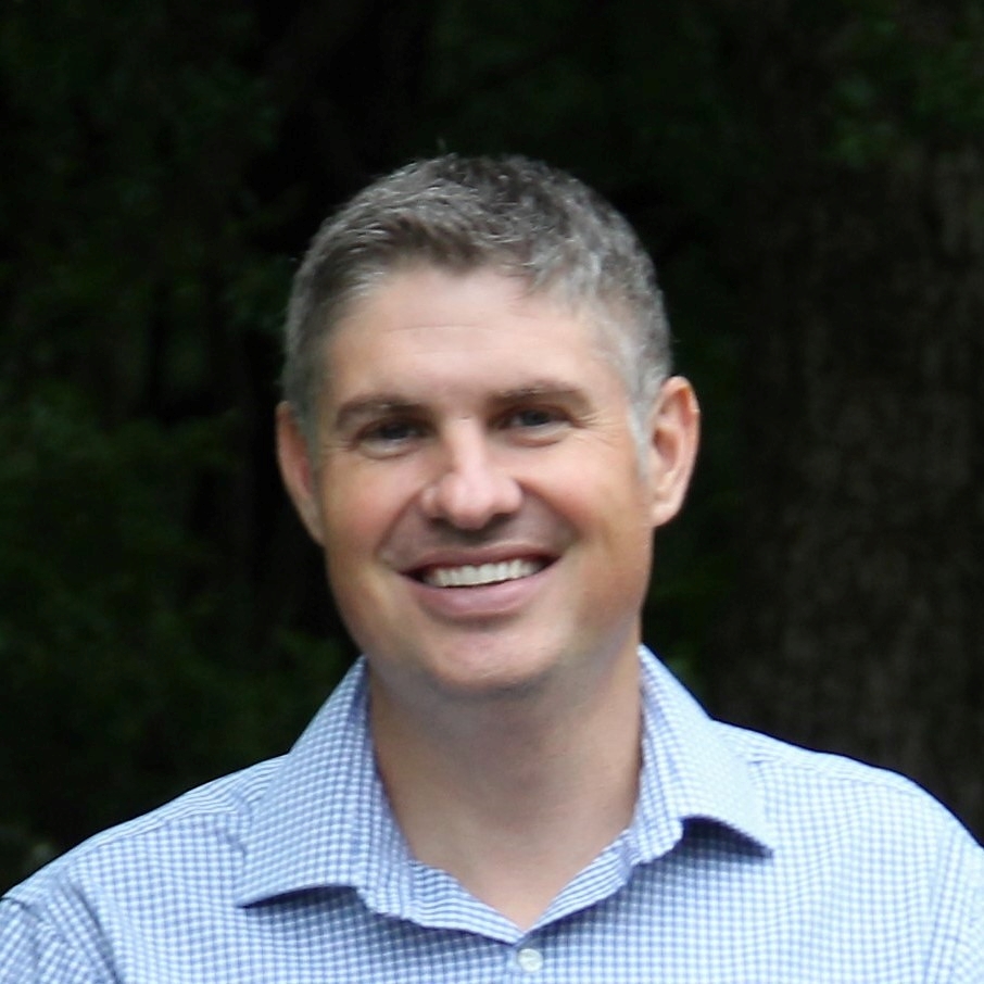 A smiling man with short gray hair wearing a blue checkered shirt outdoors with trees in the background.