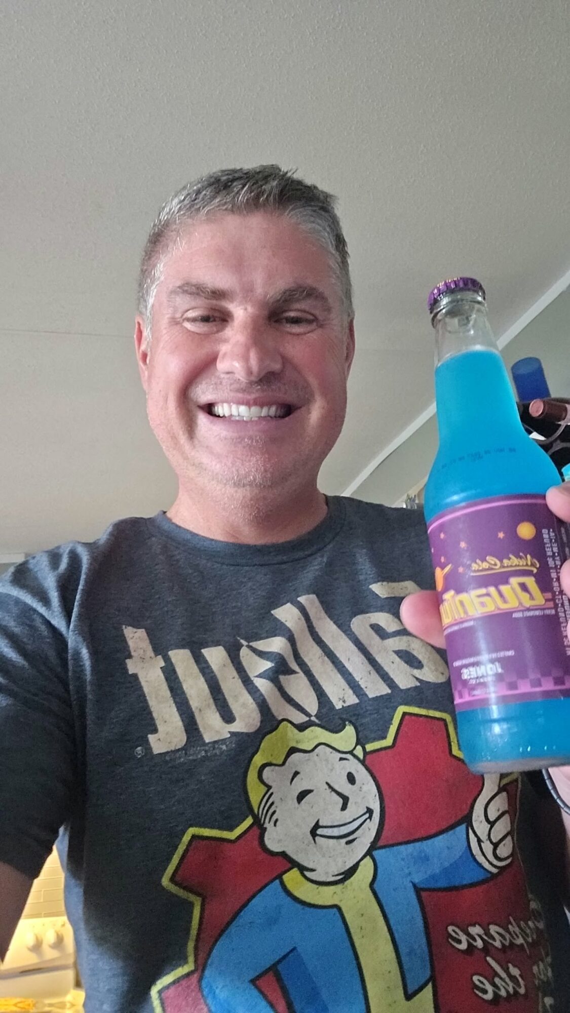 Smiling man holding a colorful bottle, wearing a graphic T-shirt, indoors with a ceiling and wall visible