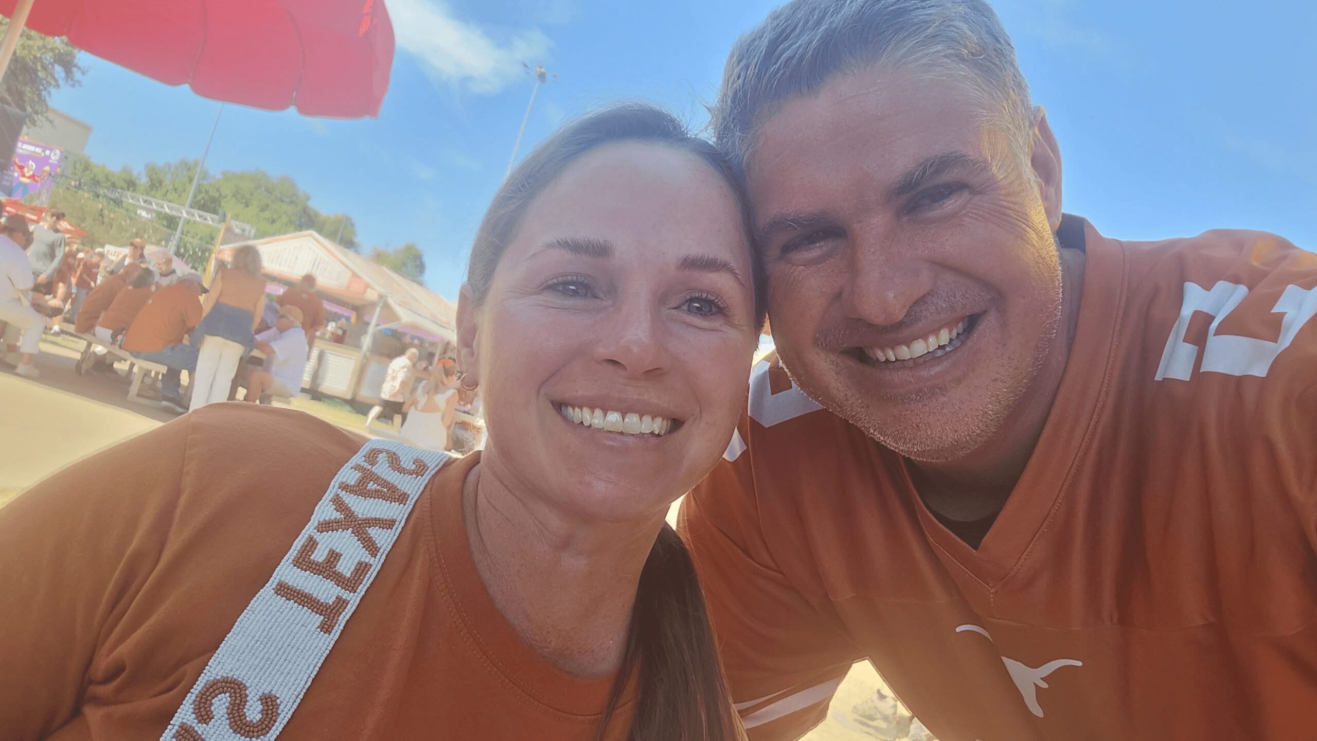 Two smiling people in orange shirts taking a selfie outdoors with tents and people in the background.