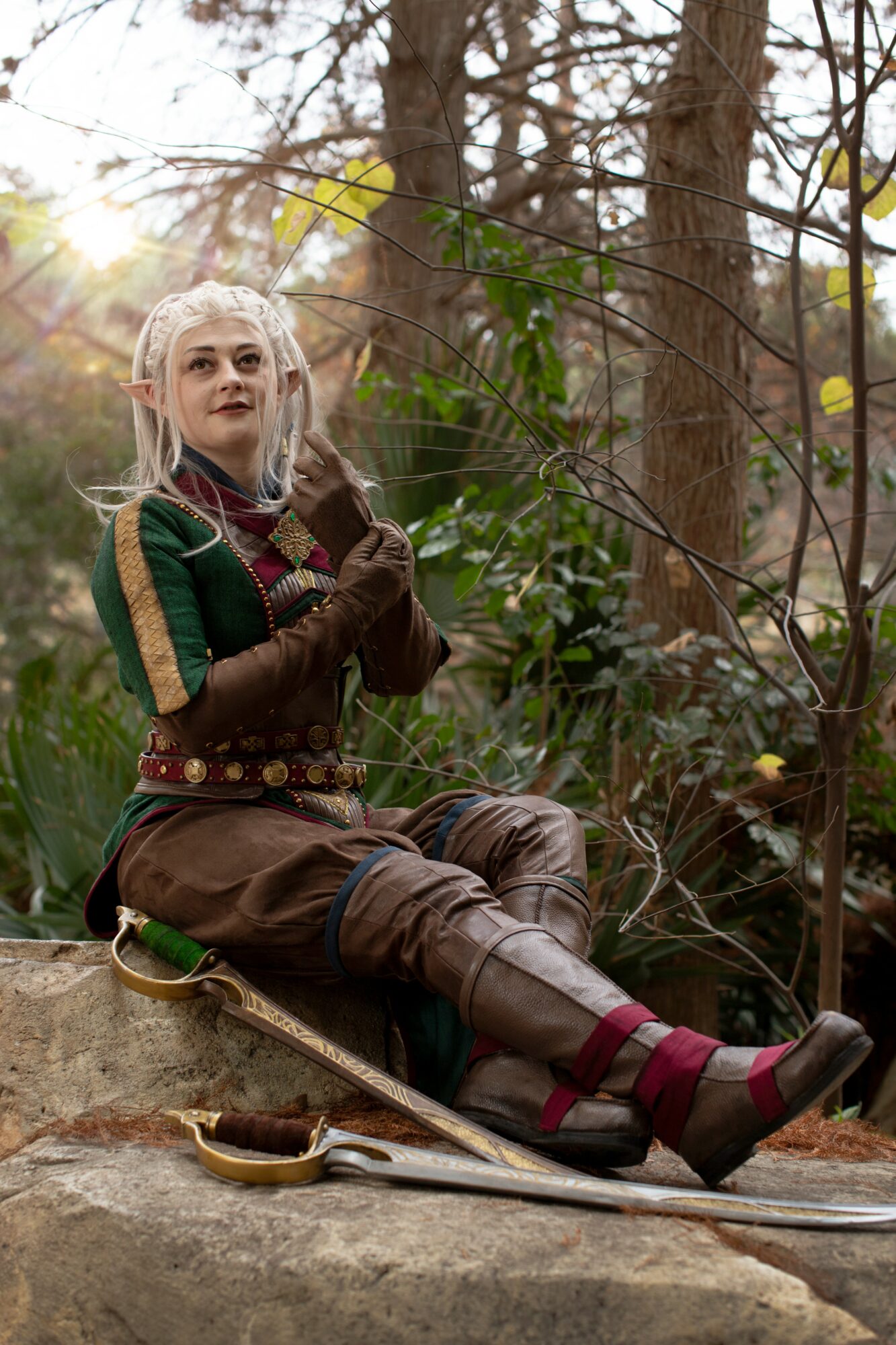 Person dressed as a fantasy character sitting on a rock in a forest, holding a bow, with trees and sunlight in background.