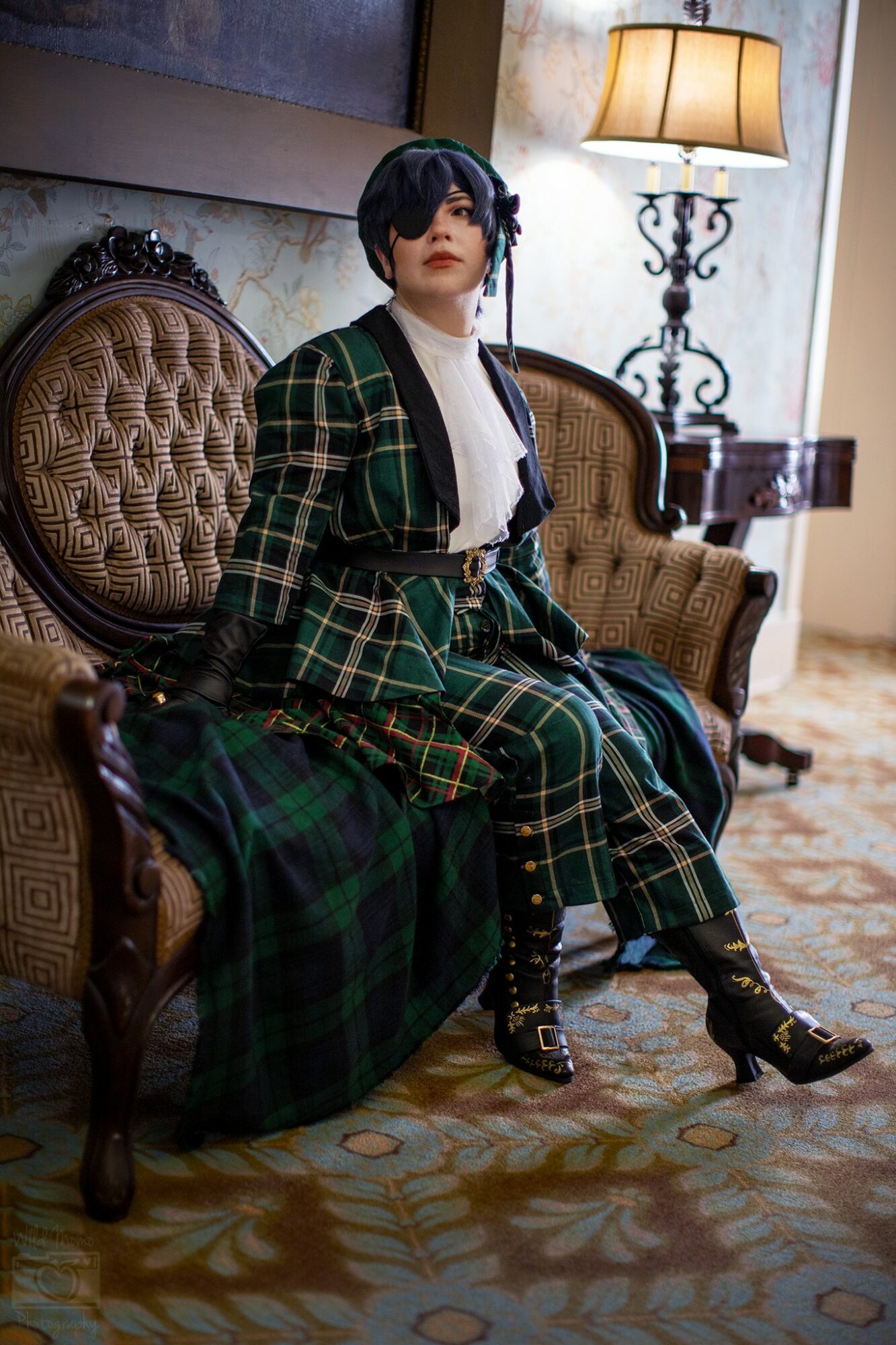 Person with short dark hair and makeup sitting on a vintage sofa in a room with a lamp and patterned carpet.