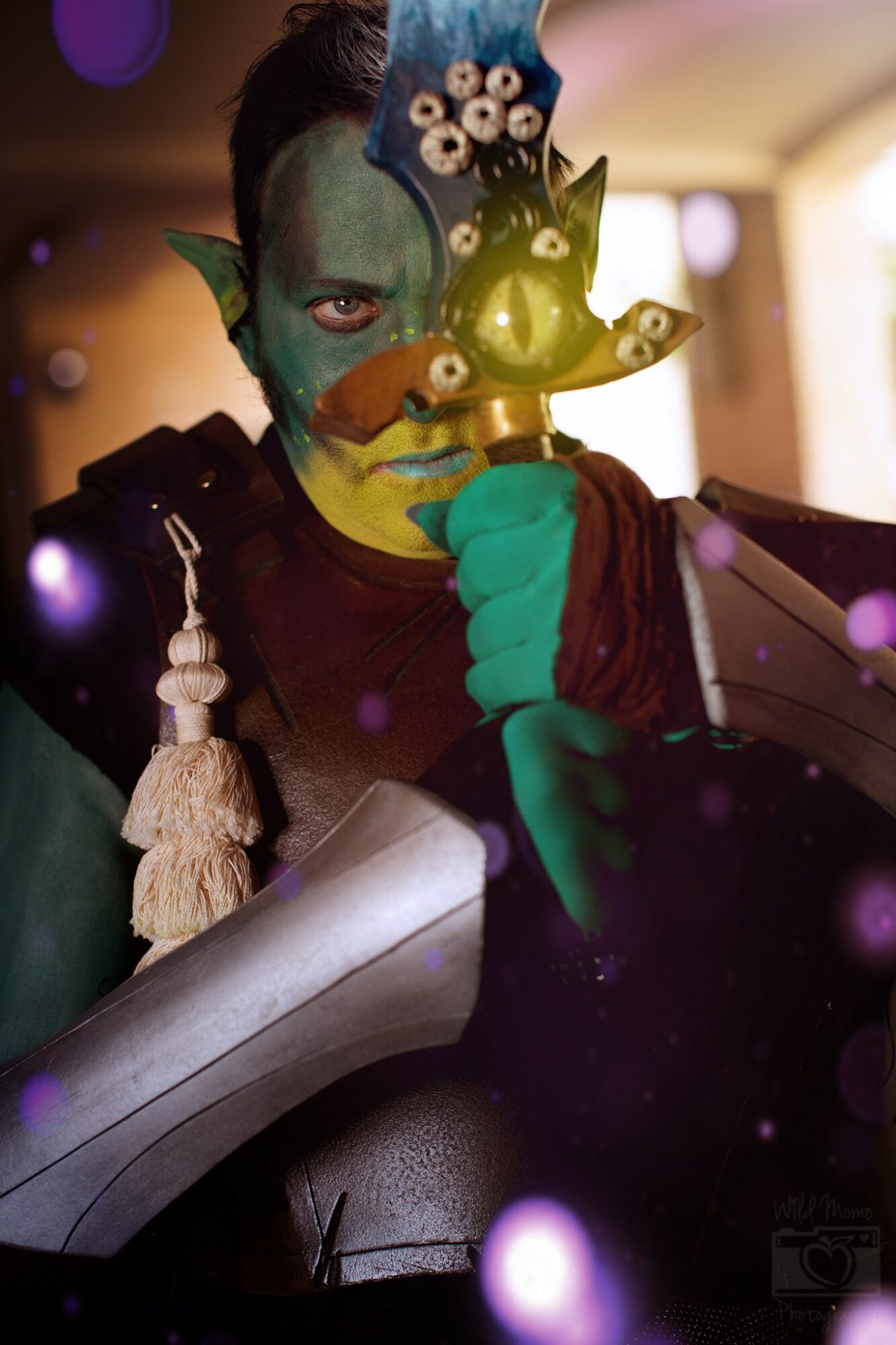 Person with colorful face paint holding a yellow pig-shaped object, wearing a costume with a tassel, in a decorated indoor setting.