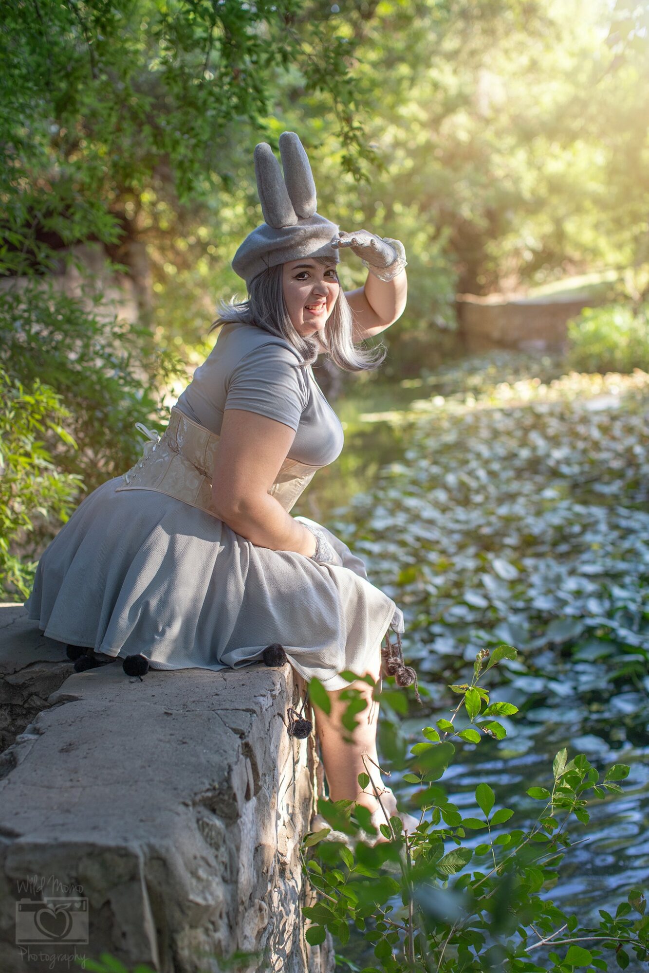 Woman sitting on a stone wall by a river, wearing a bunny hat and gray dress, with trees and sunlight in background.