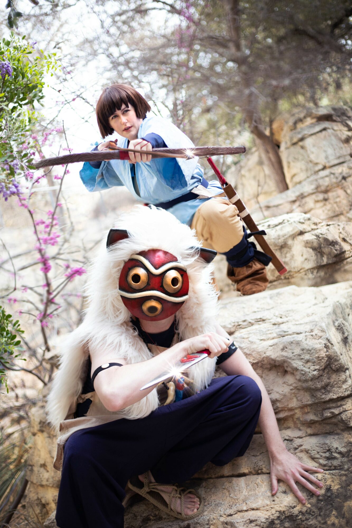 Person in a lion mask with a white mane and red, gold, and black face paint, sitting on rocks outdoors.