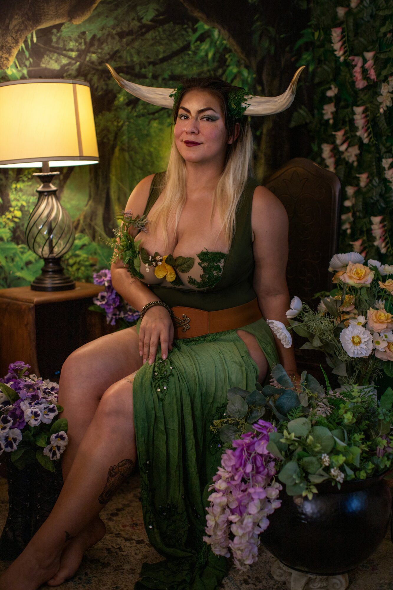 Woman dressed as a fantasy character with horns, sitting among flowers in a lush setting, illuminated by a lamp.