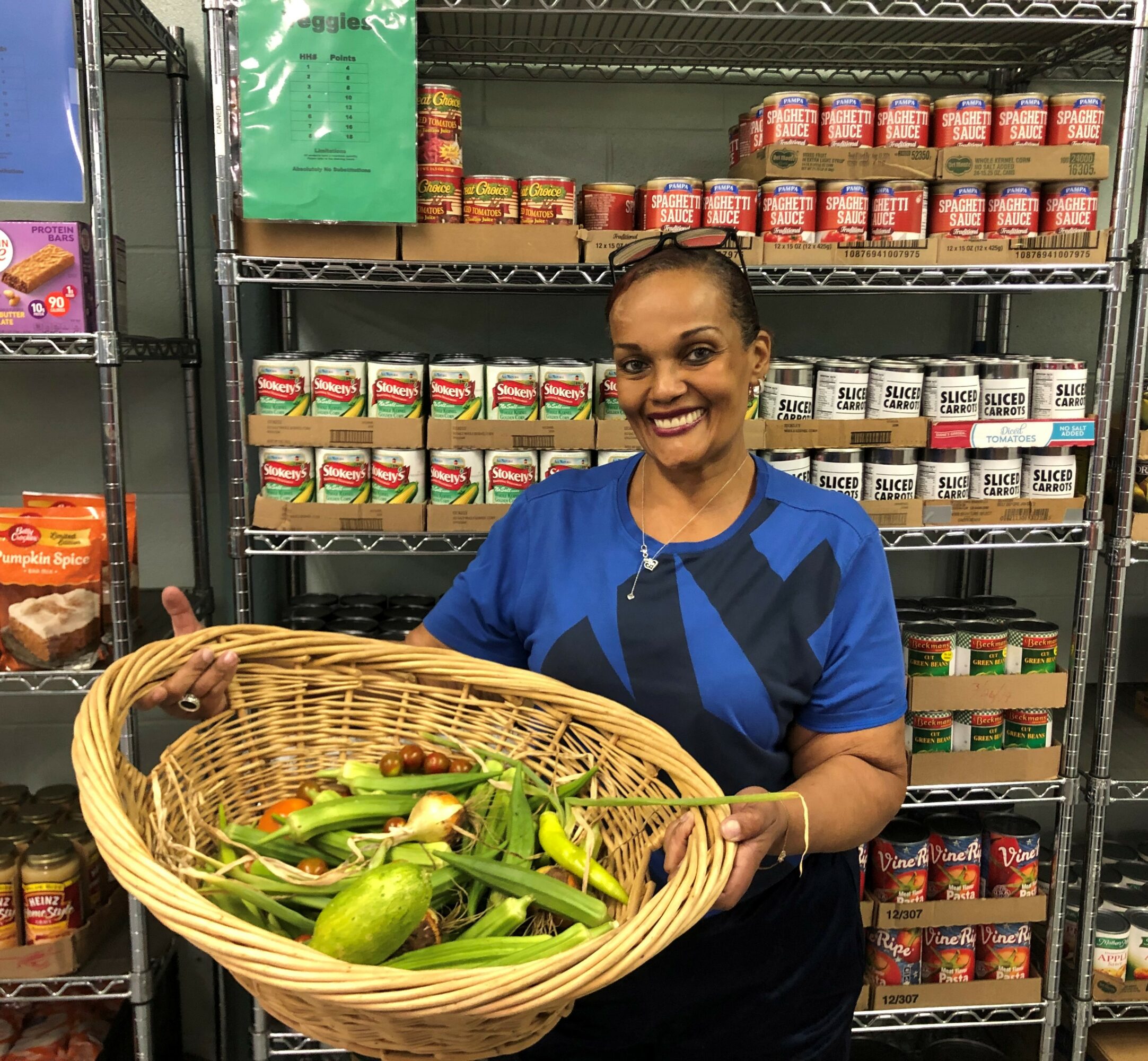 Woman holding a basket of zucchinis in a grocery store aisle with shelves of canned goods behind her.