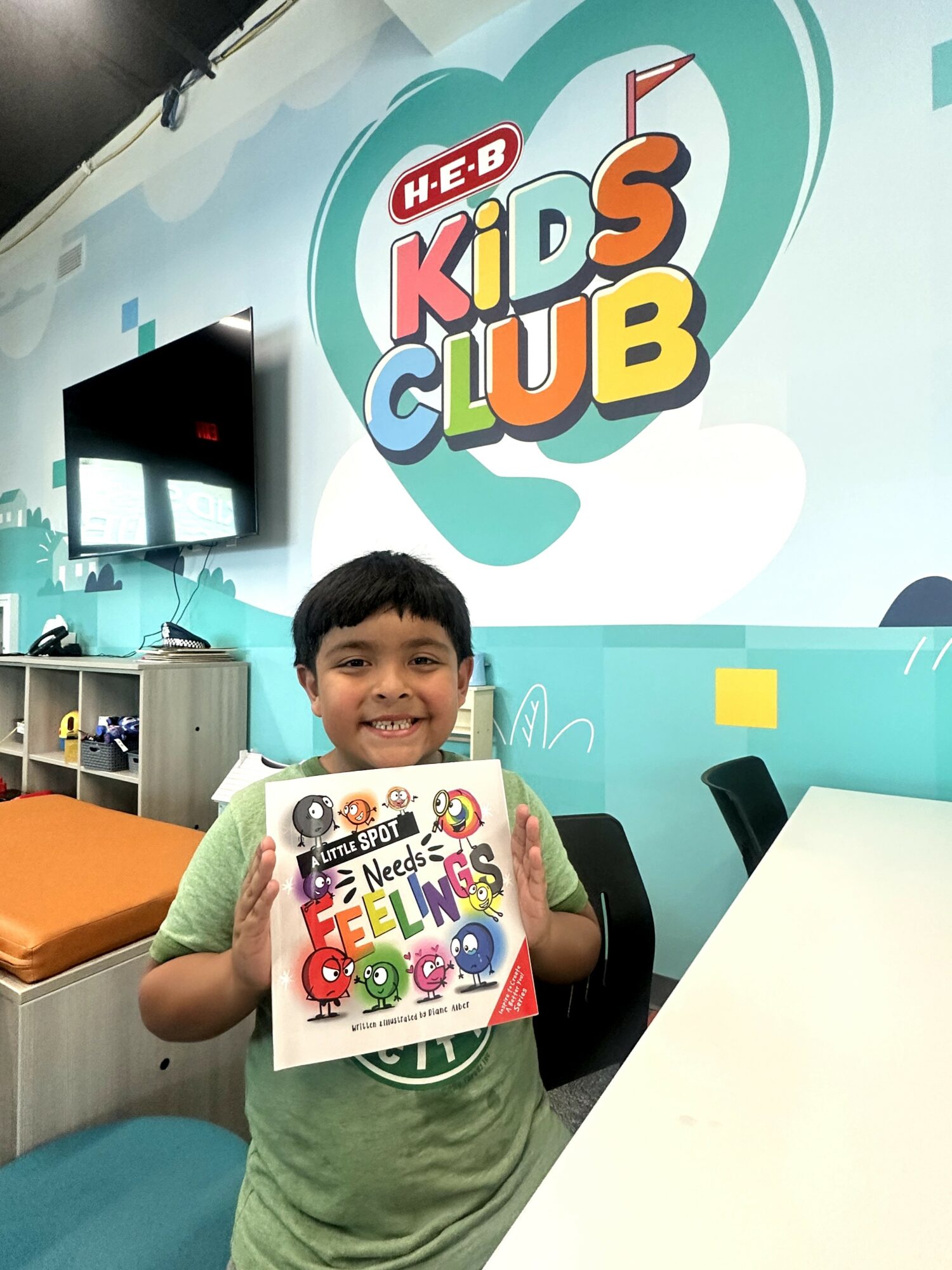 Child smiling and holding a colorful book in a kids' club room with a large H-E-B Kids Club sign on the wall.
