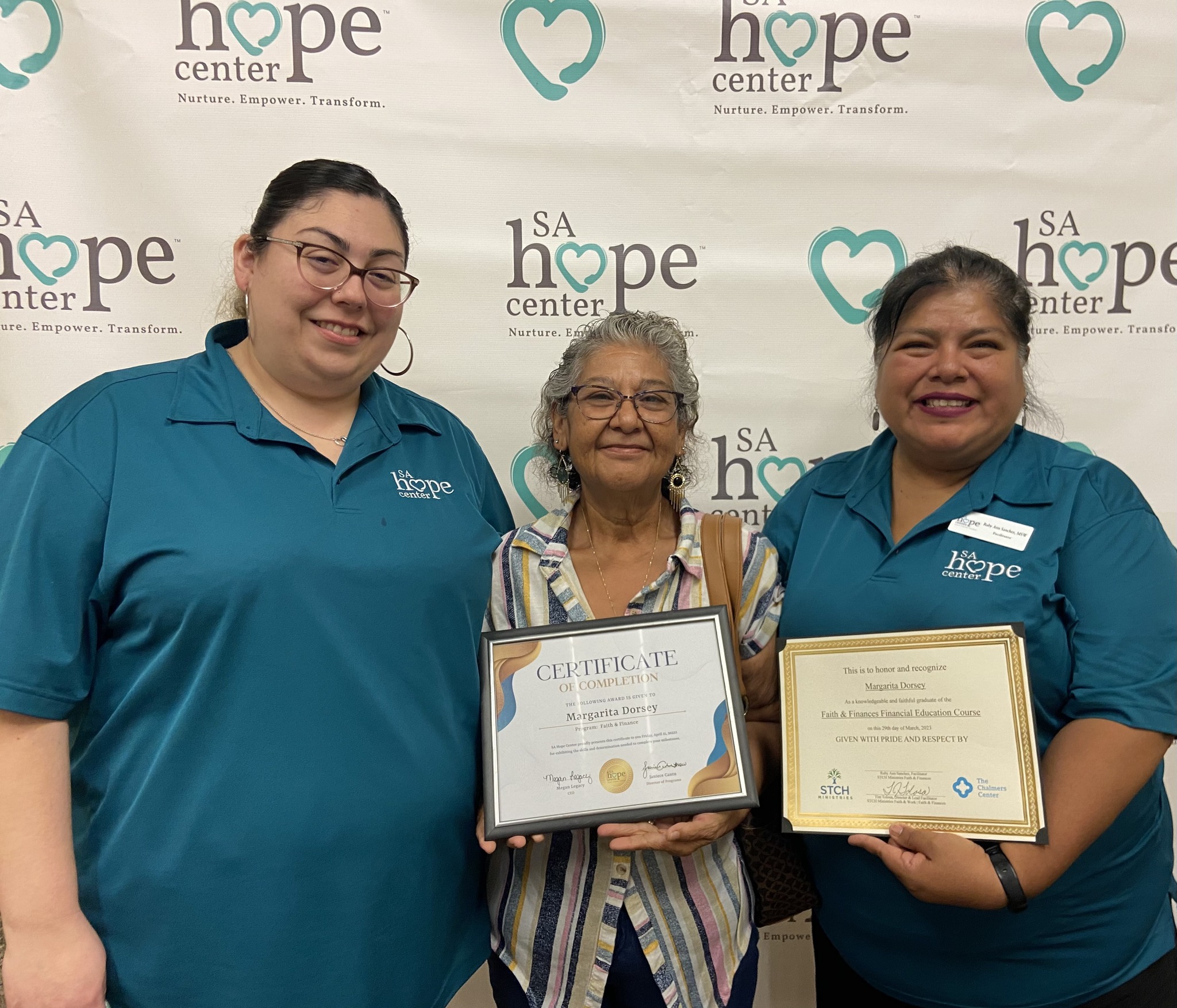Three women standing together, holding certificates, smiling, in front of a backdrop with hope center logos.