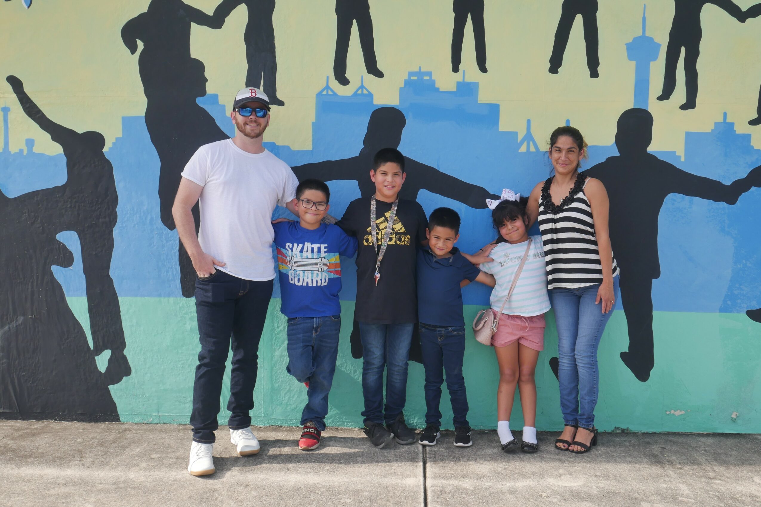 Group of six people standing in front of a colorful mural with silhouettes of people and cityscape, smiling and posing.
