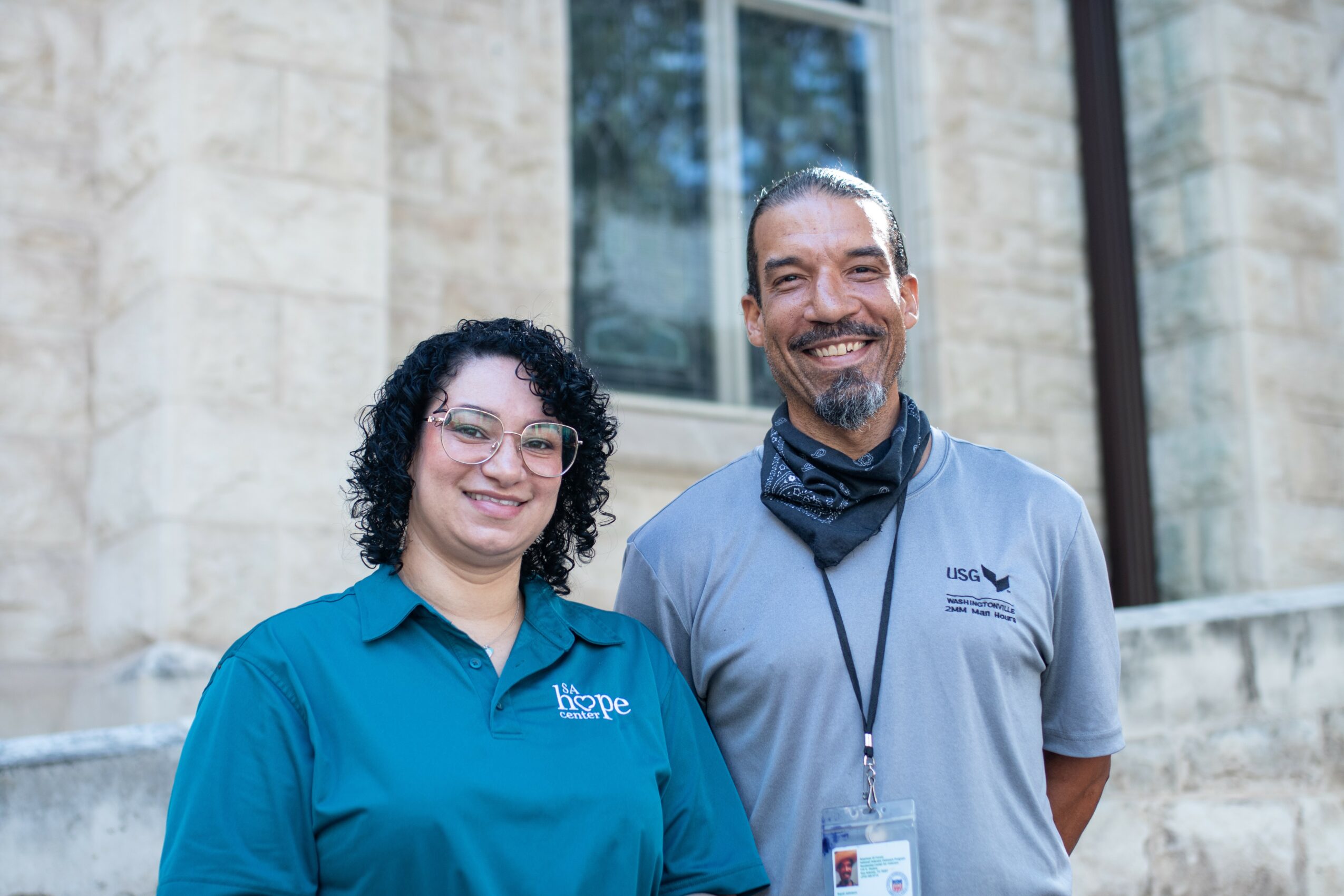 Two people standing outside in front of a stone building, smiling, one wearing a blue shirt and the other a gray shirt with a lanyard.