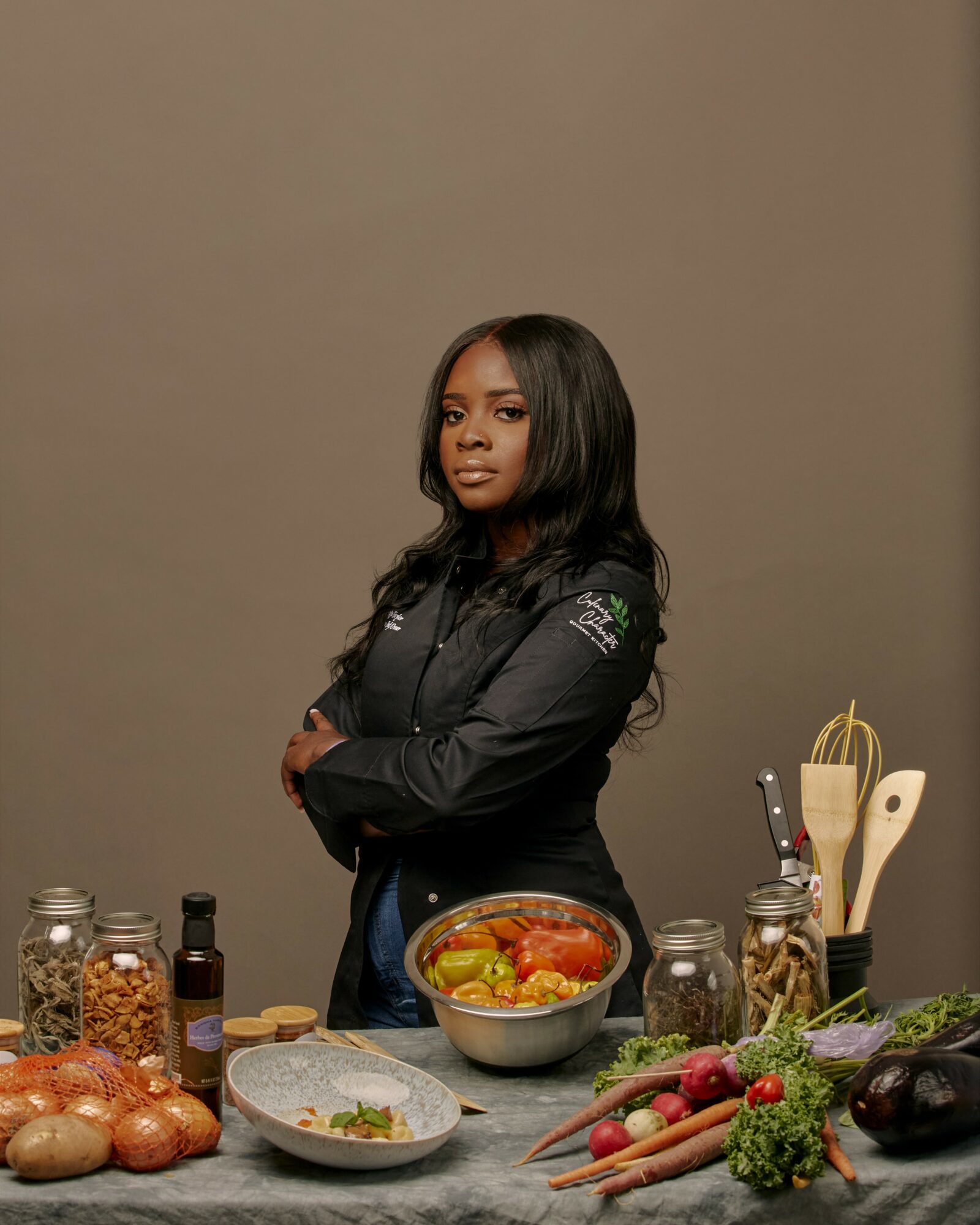 Woman in black jacket standing with arms crossed, surrounded by vegetables and kitchen utensils, against a plain background.
