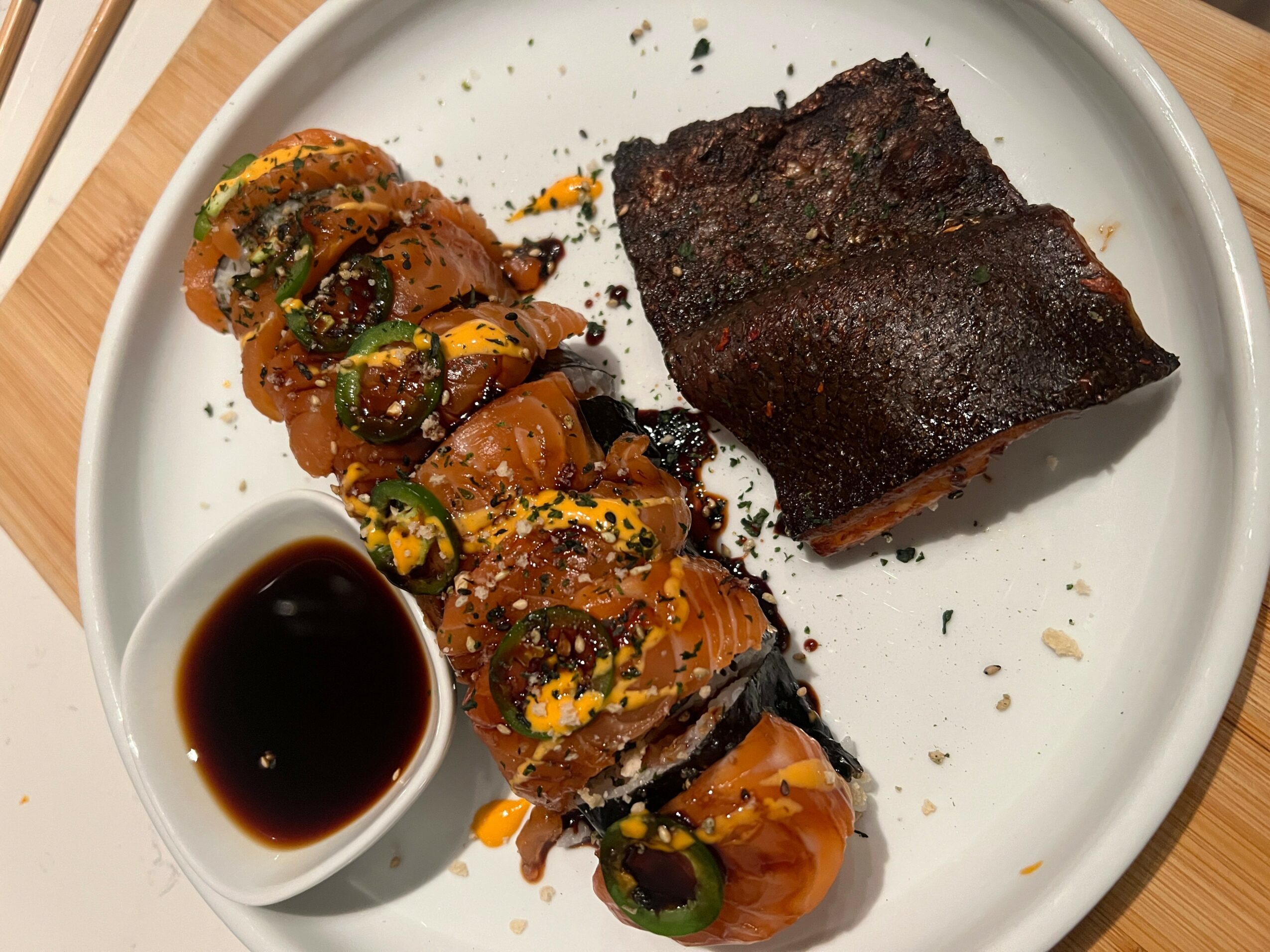 Sliced sushi roll with fish, green onions, and sauce, alongside a piece of cooked meat on a white plate.