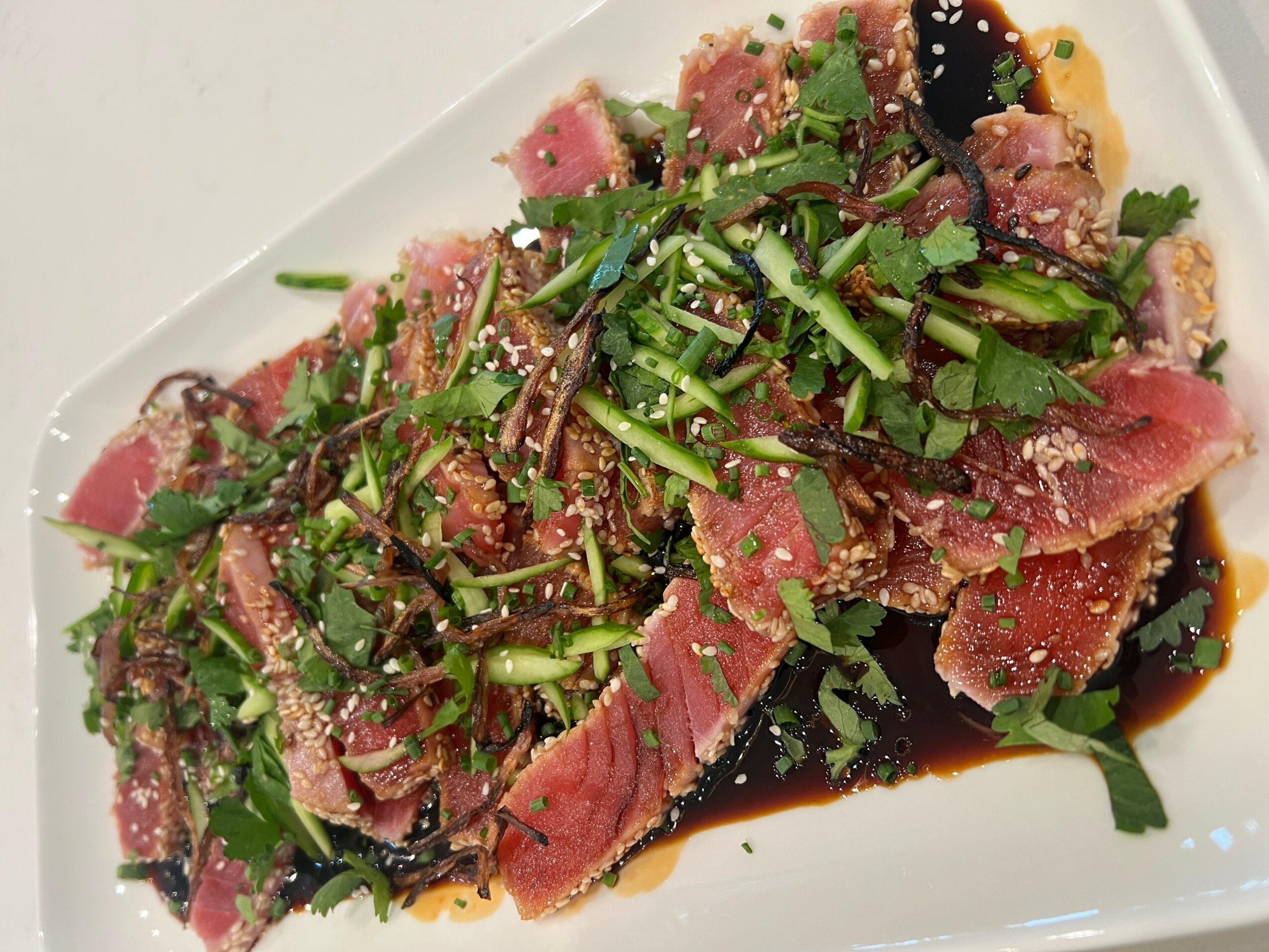 Sliced seared tuna topped with chopped green herbs and sesame seeds, drizzled with dark sauce on a white plate.