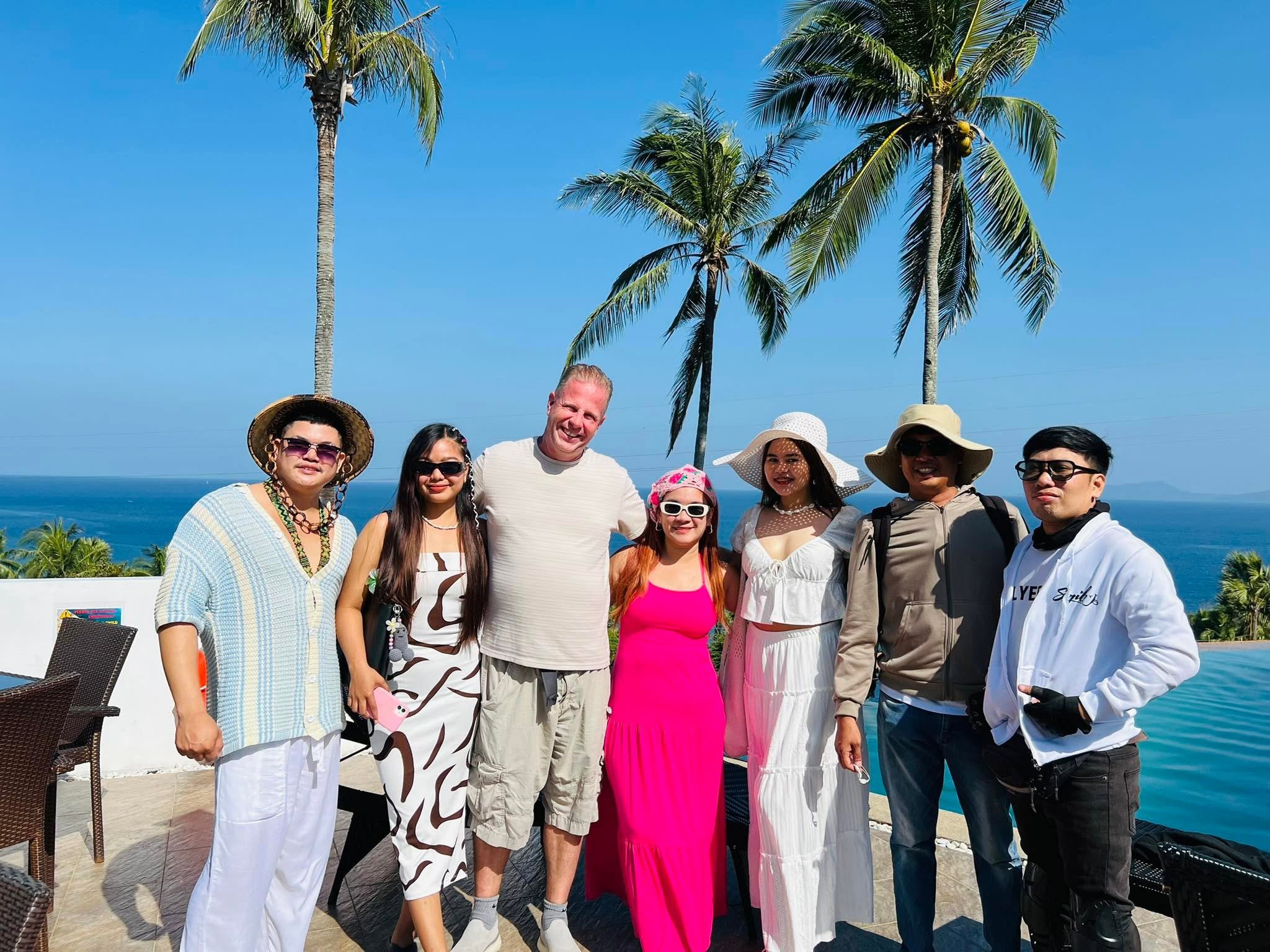 Group of seven people standing outdoors with palm trees and ocean in background, smiling, dressed in casual summer clothing.