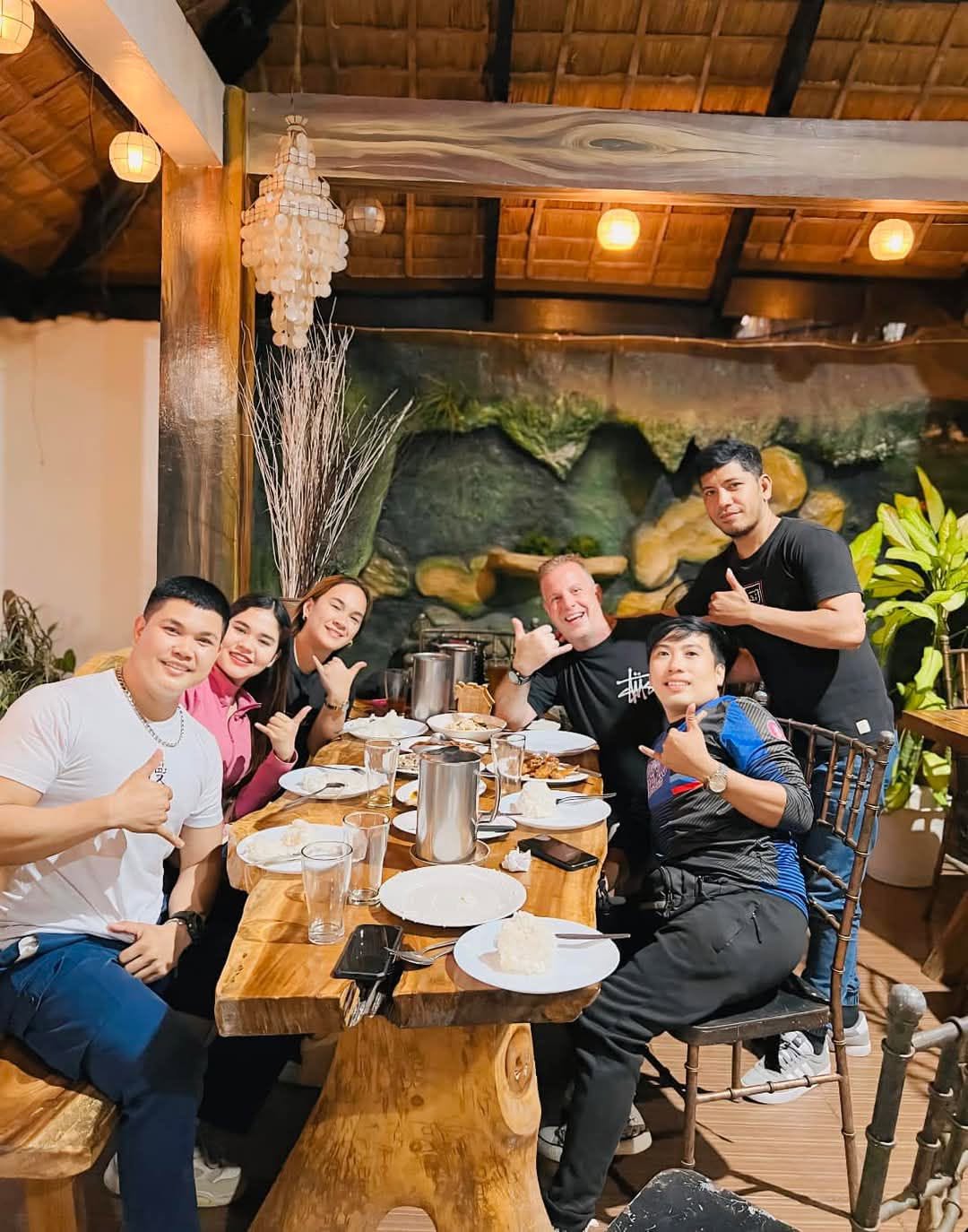 Group of seven people sitting and standing around a dining table in a restaurant, smiling and giving thumbs up, with plants and decor in background.