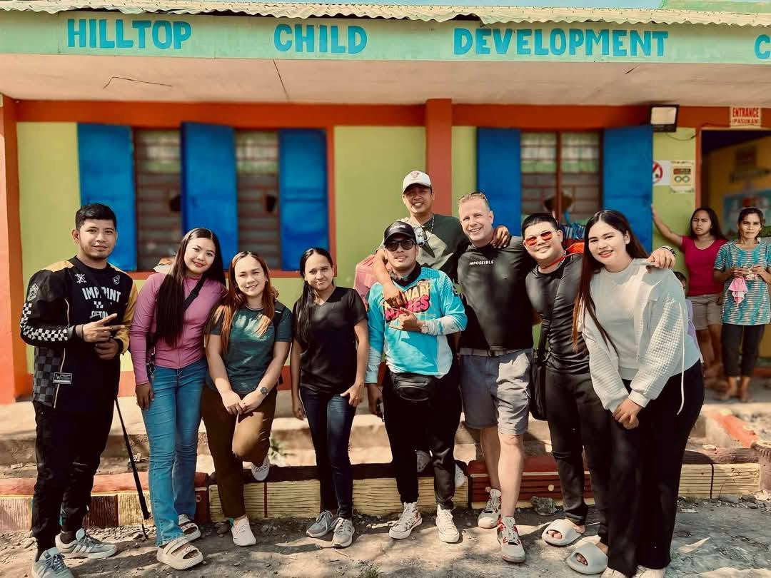 Group of nine people standing outside a building with colorful pillars, smiling, with a sign reading 'HILLTOP CHILD DEVELOPMENT' above.